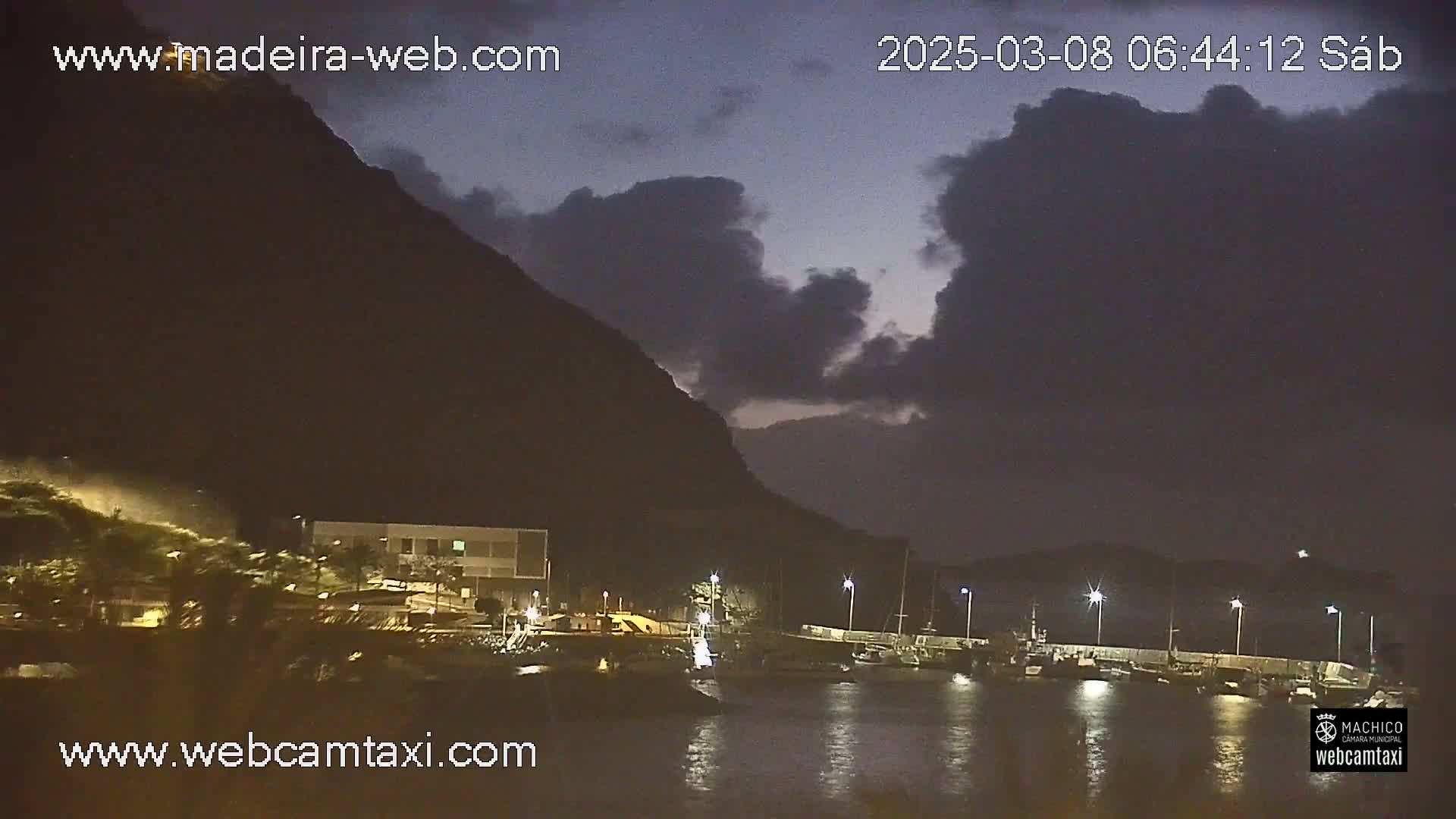 A nighttime harbor scene shows several boats moored near a dark, mountainous shore under a partly cloudy sky.
