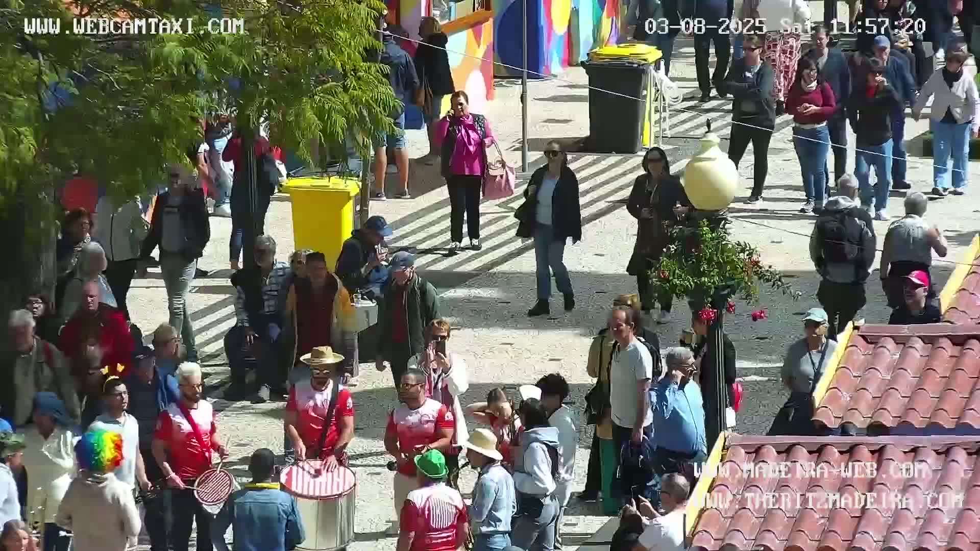 A sunny outdoor scene shows a crowd of people milling about a paved plaza, some playing drums, others standing in groups, and some walking around.