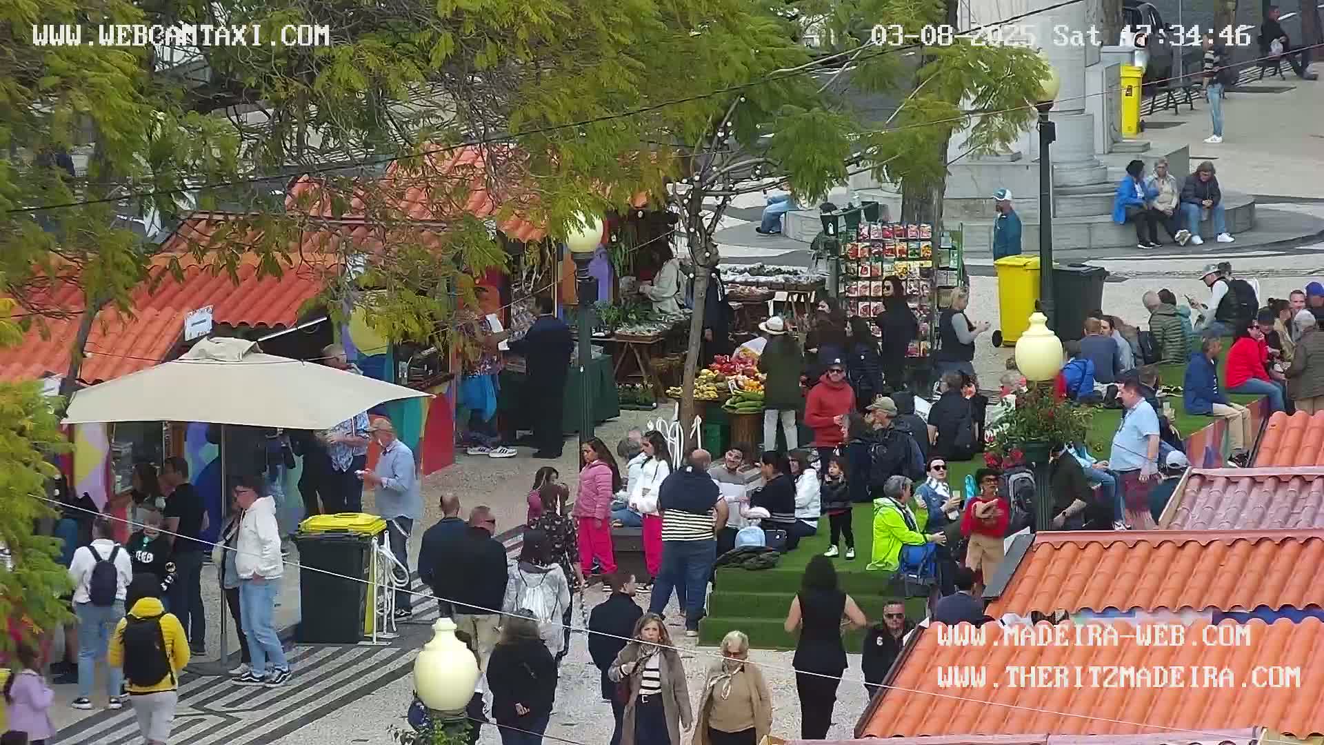 A sunny outdoor market scene with numerous people browsing stalls and sitting on a grassy area, surrounded by colorful buildings with red-tiled roofs.