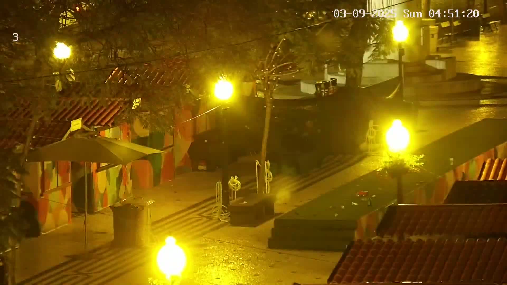 Under the yellow glow of streetlights in the rain at night, a plaza scene shows colorful buildings, an umbrella, and what appears to be a dark vehicle.
