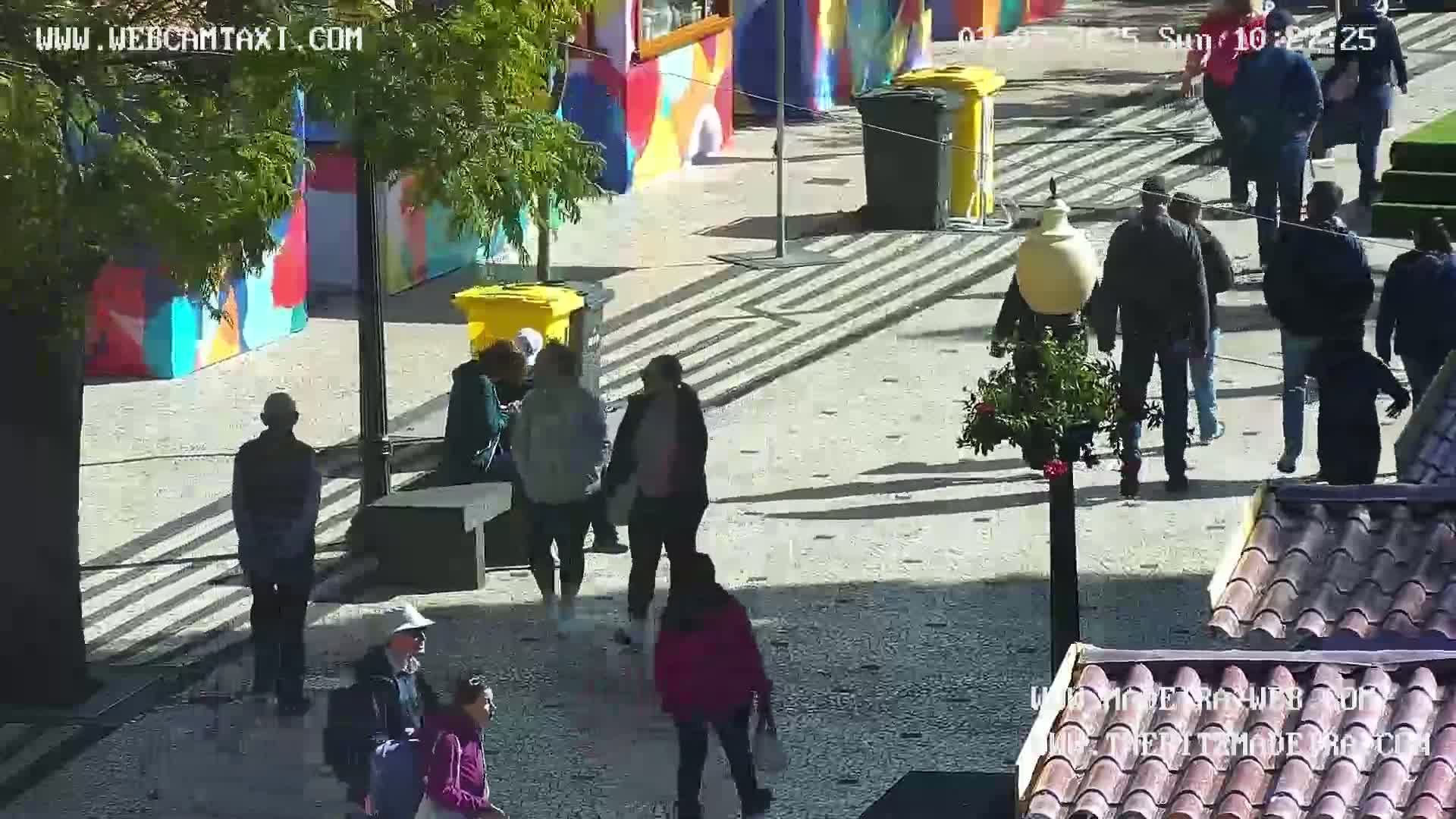 A sunny outdoor plaza with several people walking around, passing by colorful buildings and yellow trash cans.