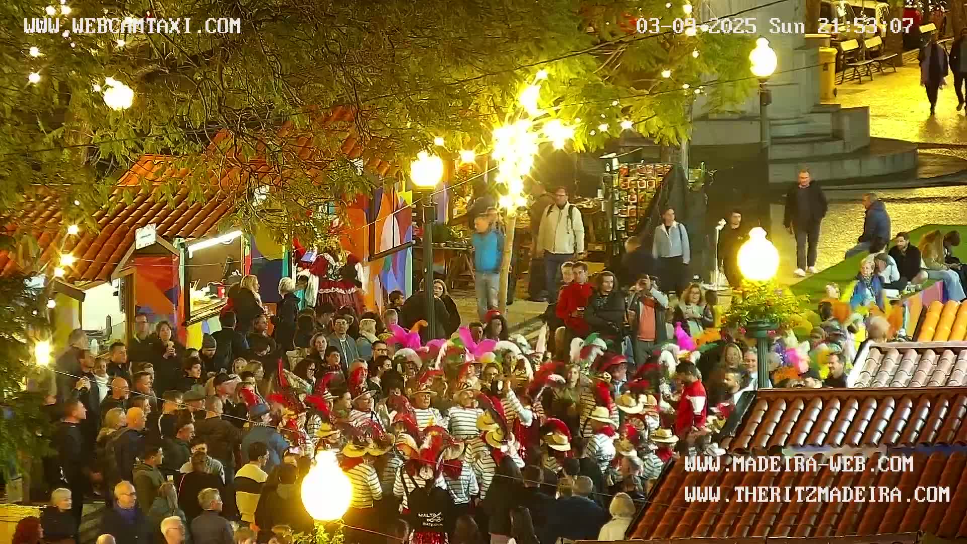 A large crowd of people, many in costume, are gathered in a brightly lit outdoor plaza at night under strings of lights.
