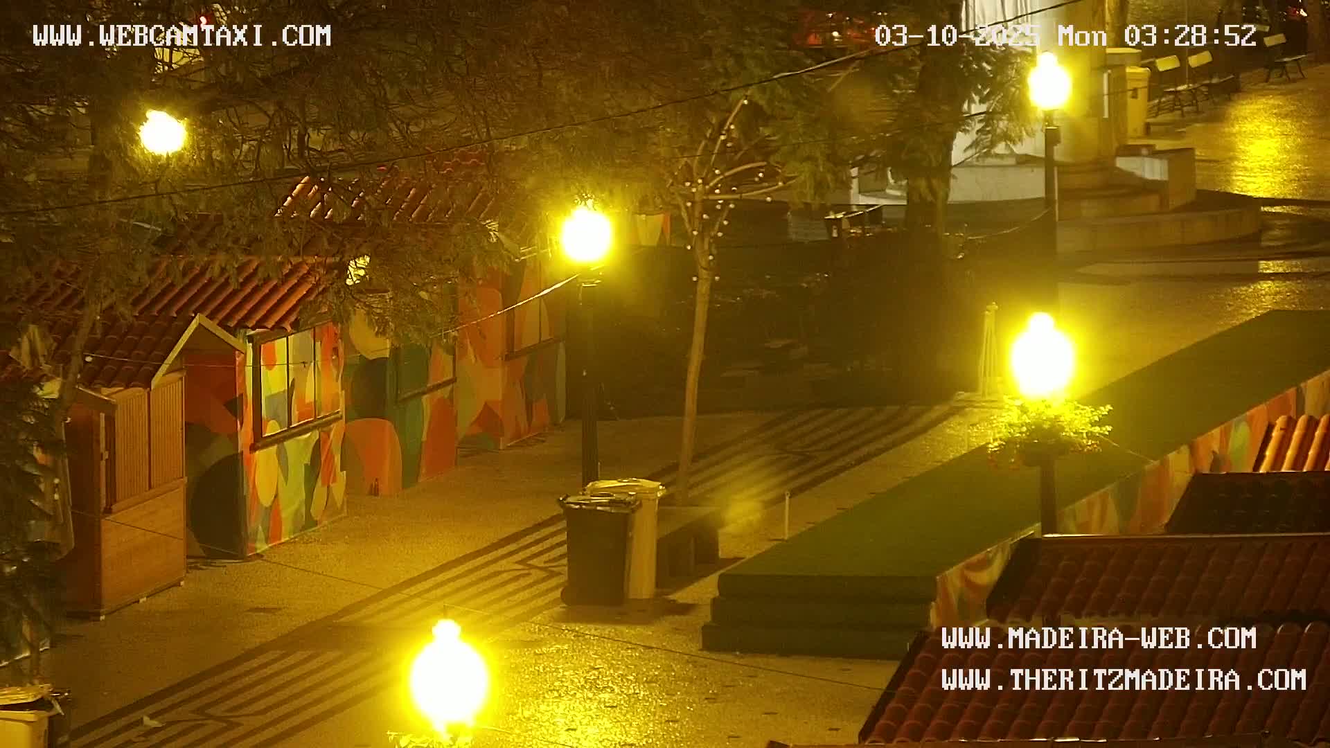 A rain-slicked, nighttime plaza scene shows brightly lit colorful kiosks and streetlamps under a dark sky.