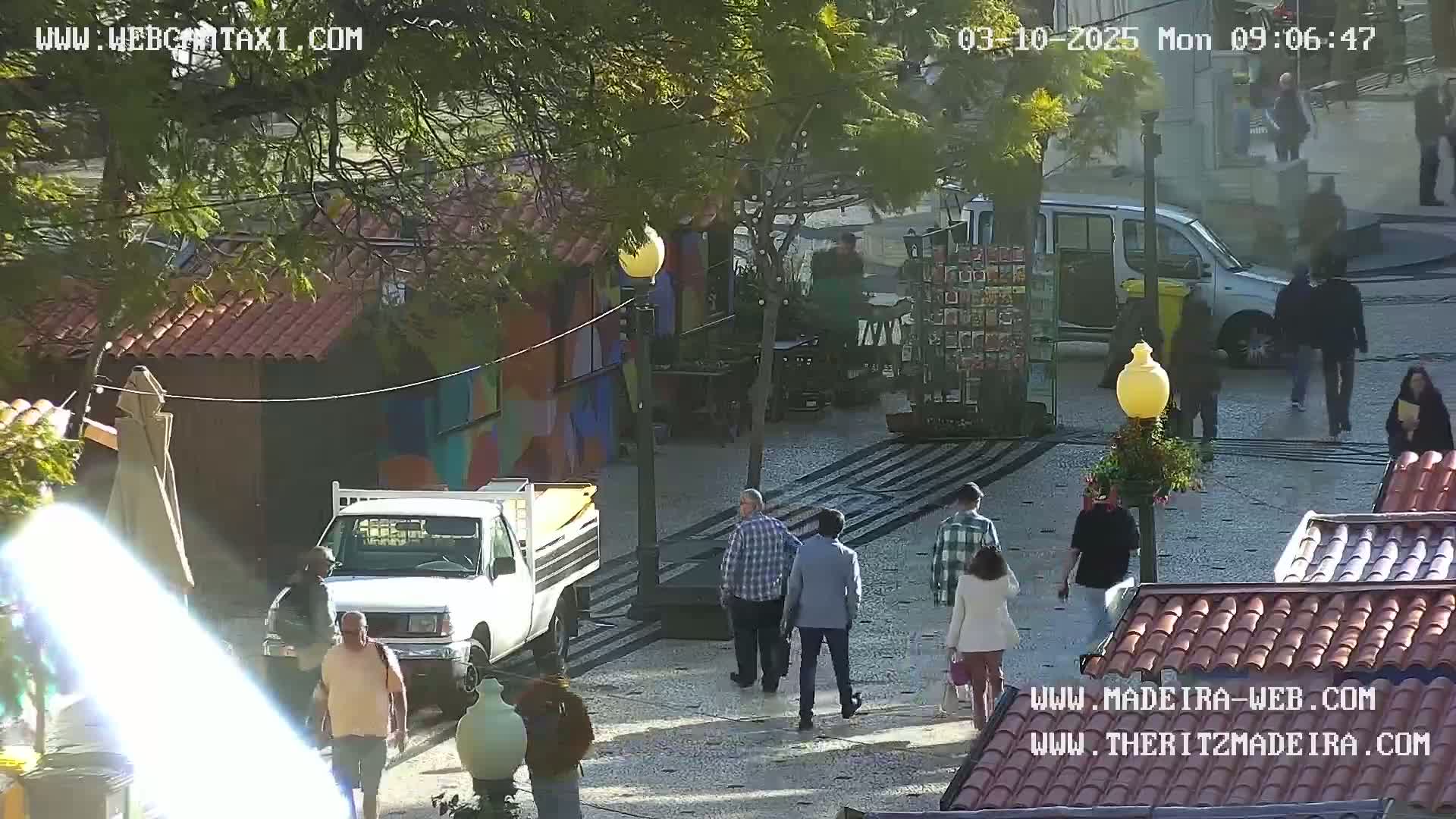 A sunny outdoor plaza scene shows several people walking amidst small market stalls and parked vehicles.