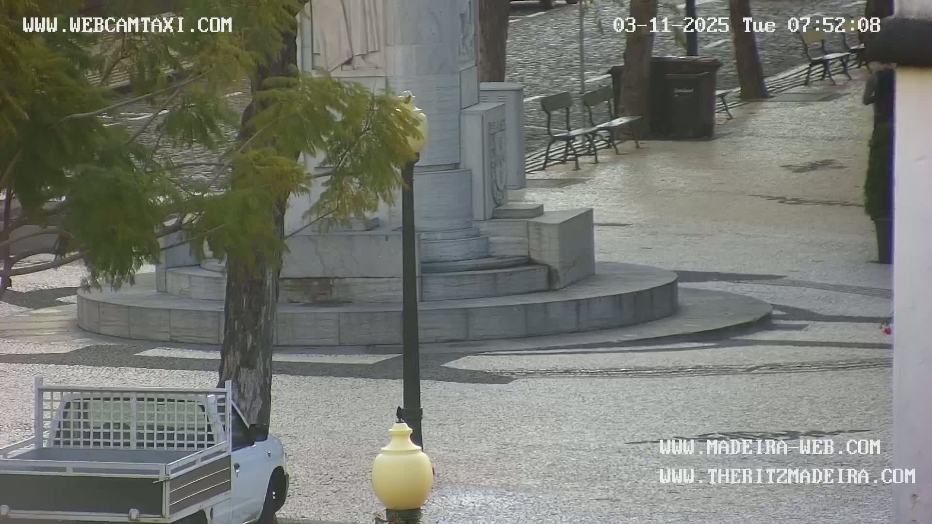 A white pickup truck is parked near a multi-tiered stone monument in a plaza on a partly cloudy day.