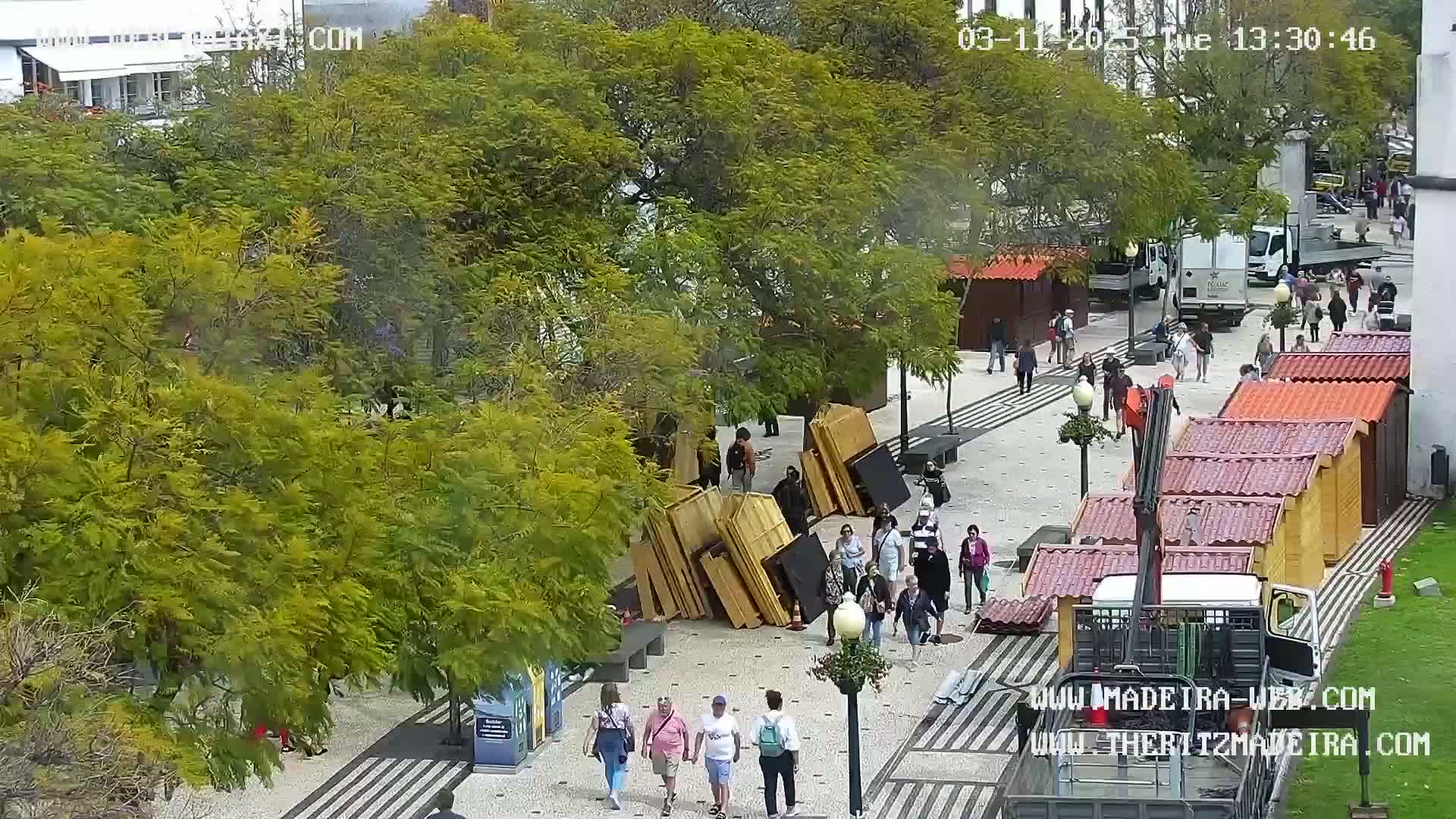 A sunny outdoor plaza scene shows numerous people walking among stacked wooden structures and small market stalls, with a crane truck parked nearby.