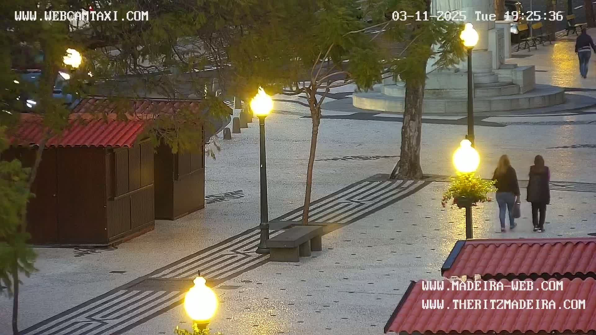 Two women walk across a stone plaza at dusk, past illuminated lampposts and a small kiosk, under a clear sky.