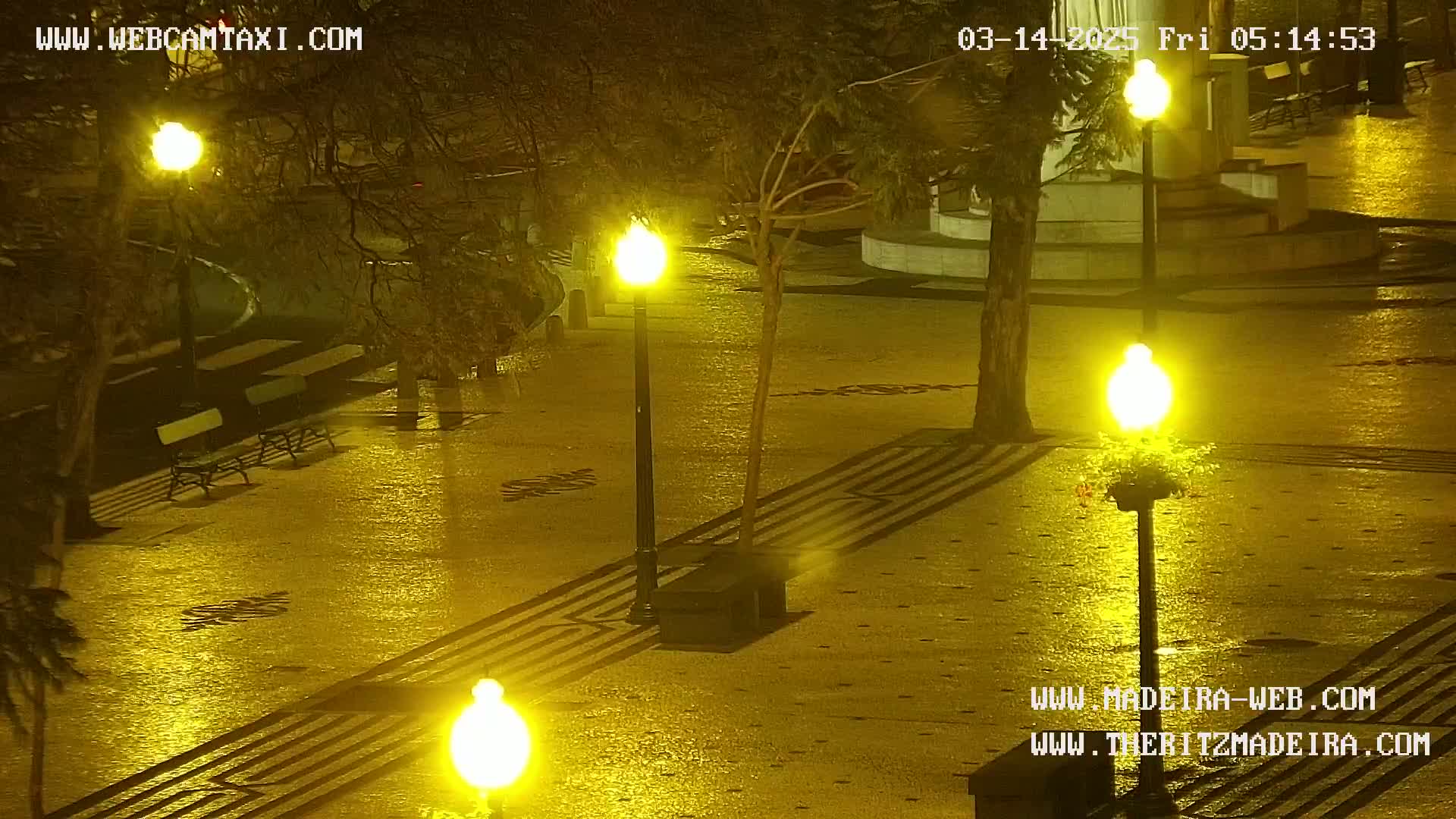 A wet, nighttime paved plaza is illuminated by several bright streetlights, with empty benches visible in the background.
