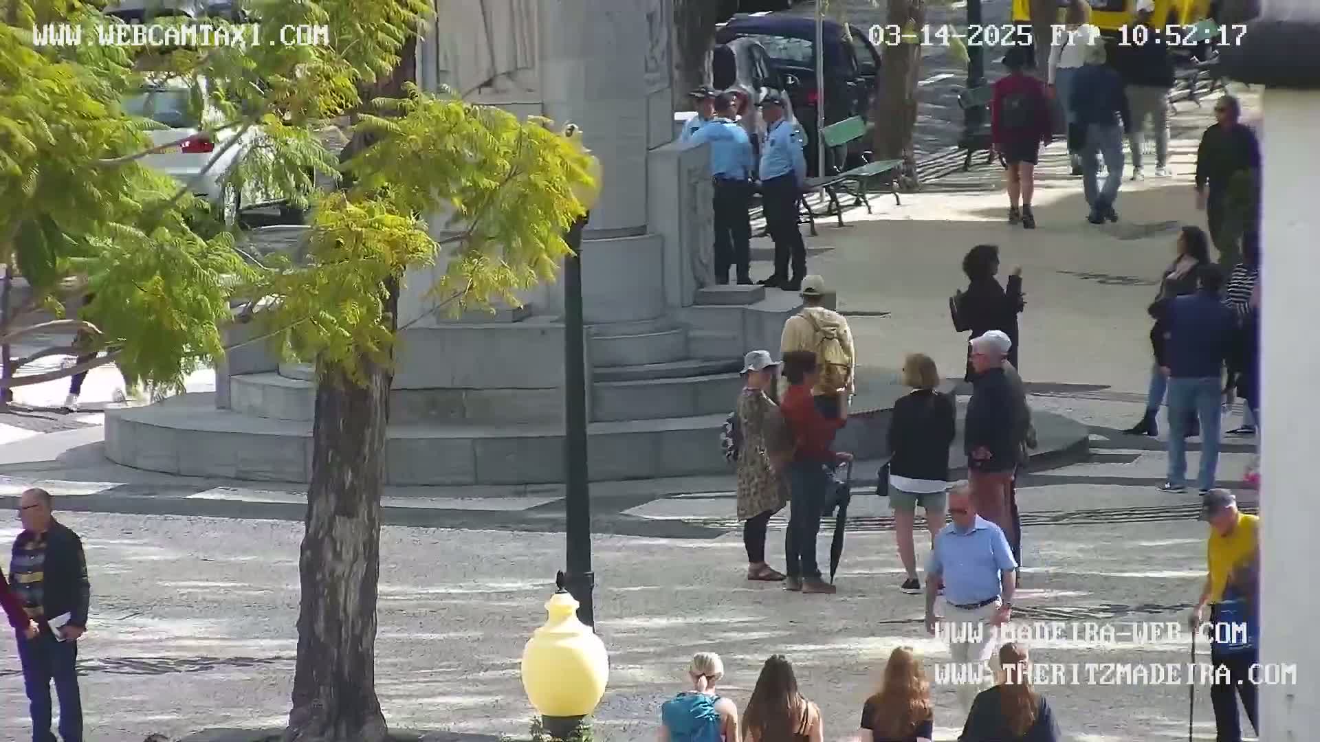 A sunny day in a plaza with several people walking around near a stone monument and a tree.
