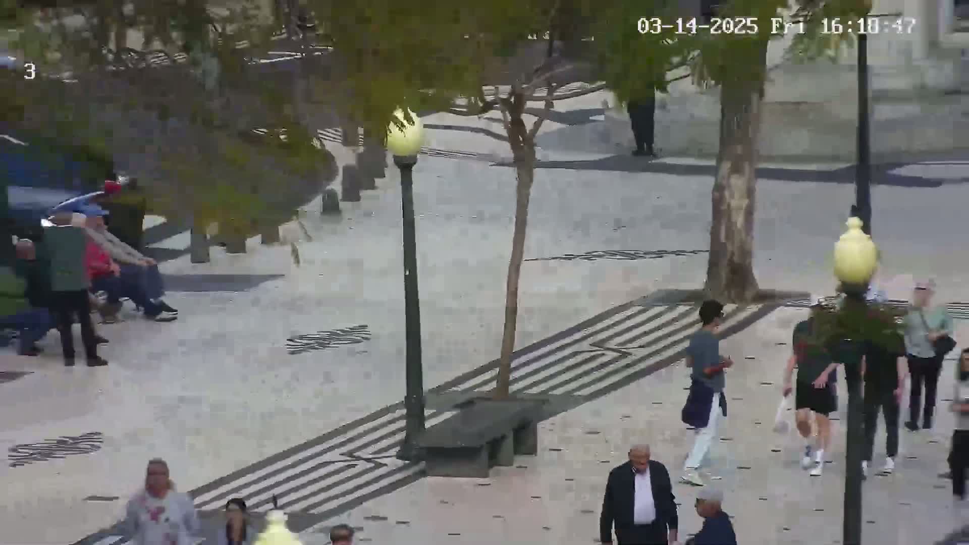 A paved pedestrian area with several people walking, some sitting, and trees with light poles, under a sunny sky.