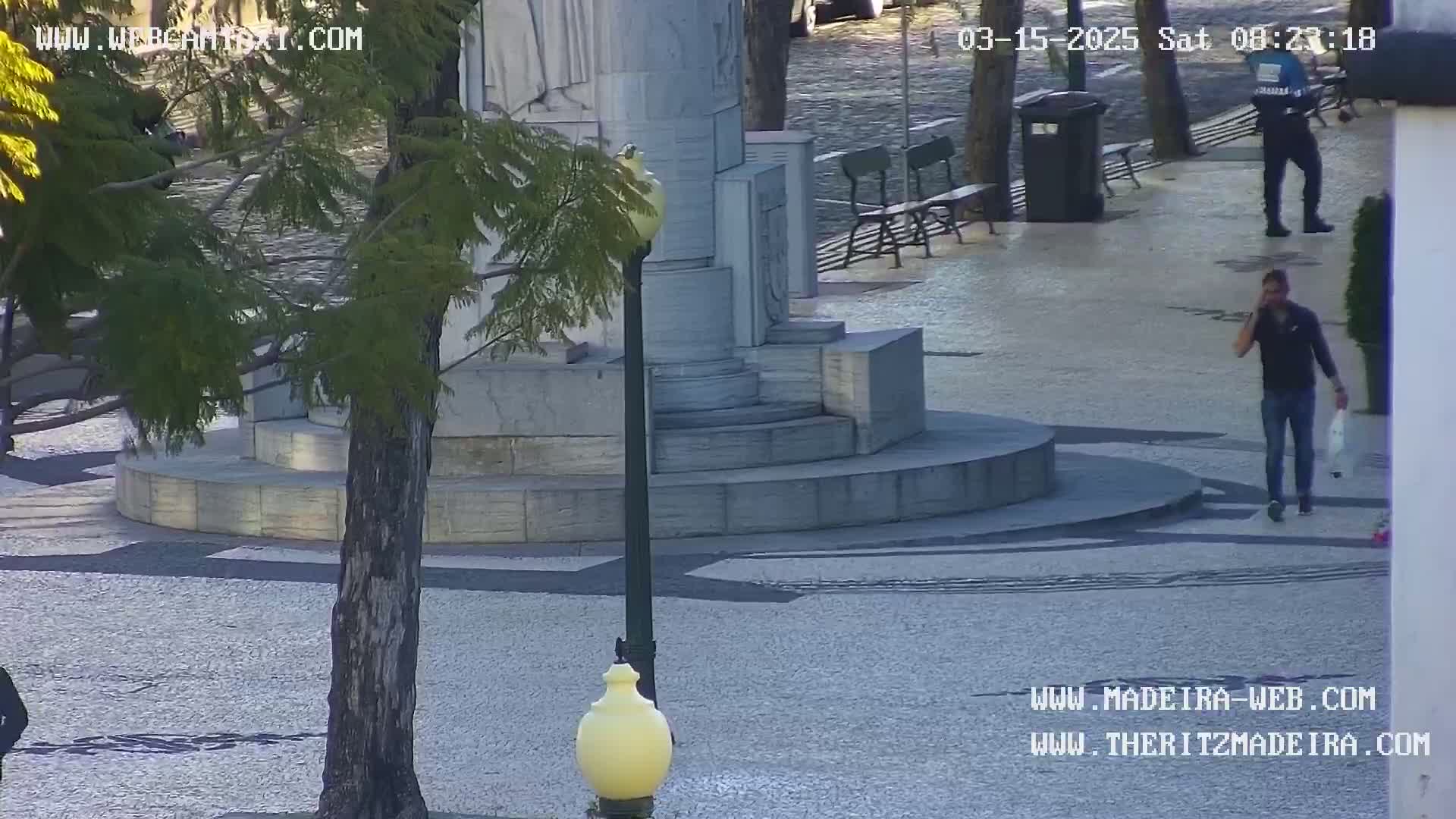 A man on a cell phone walks past a stone monument and several benches in a sunny, paved plaza.
