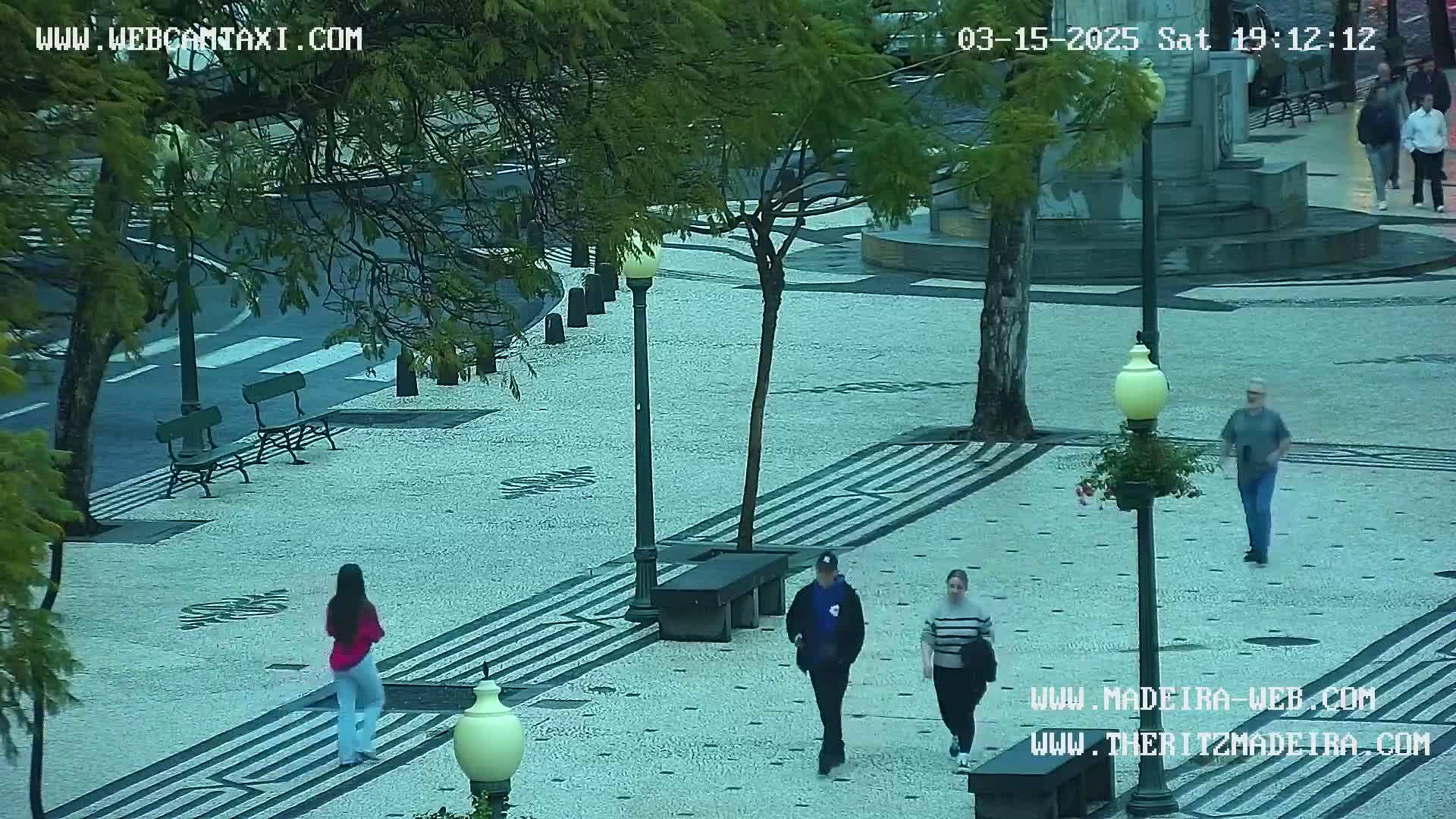 A wet, paved plaza with several people walking, some benches, and trees under a cloudy sky.