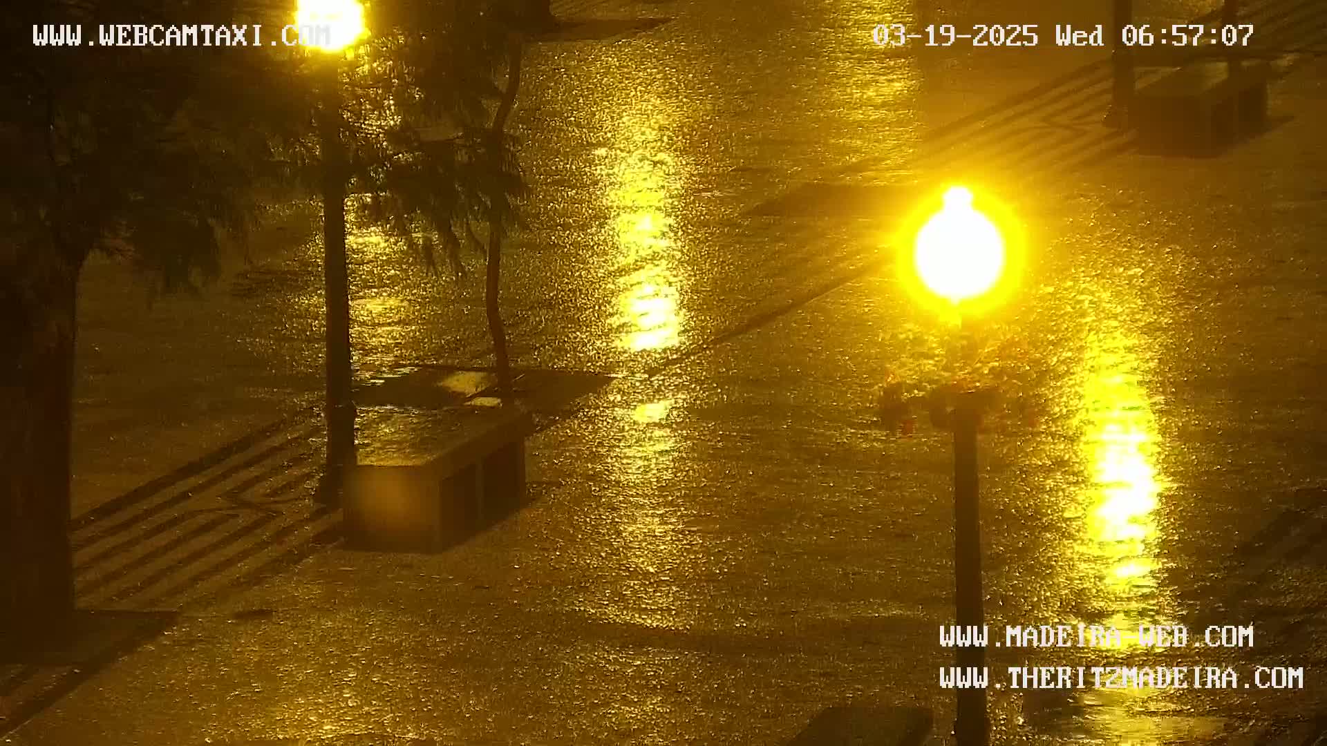 A rain-soaked street at night is illuminated by bright streetlights, reflecting on the flooded pavement.