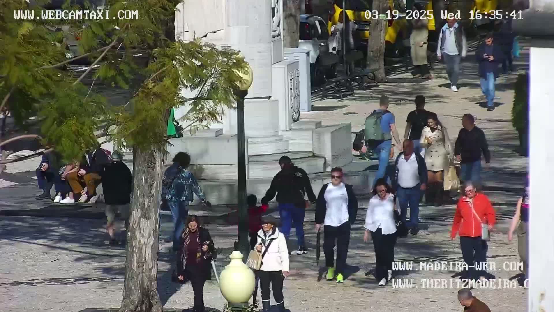 A sunny outdoor plaza scene shows many people walking and sitting around a monument, some in groups and some alone.