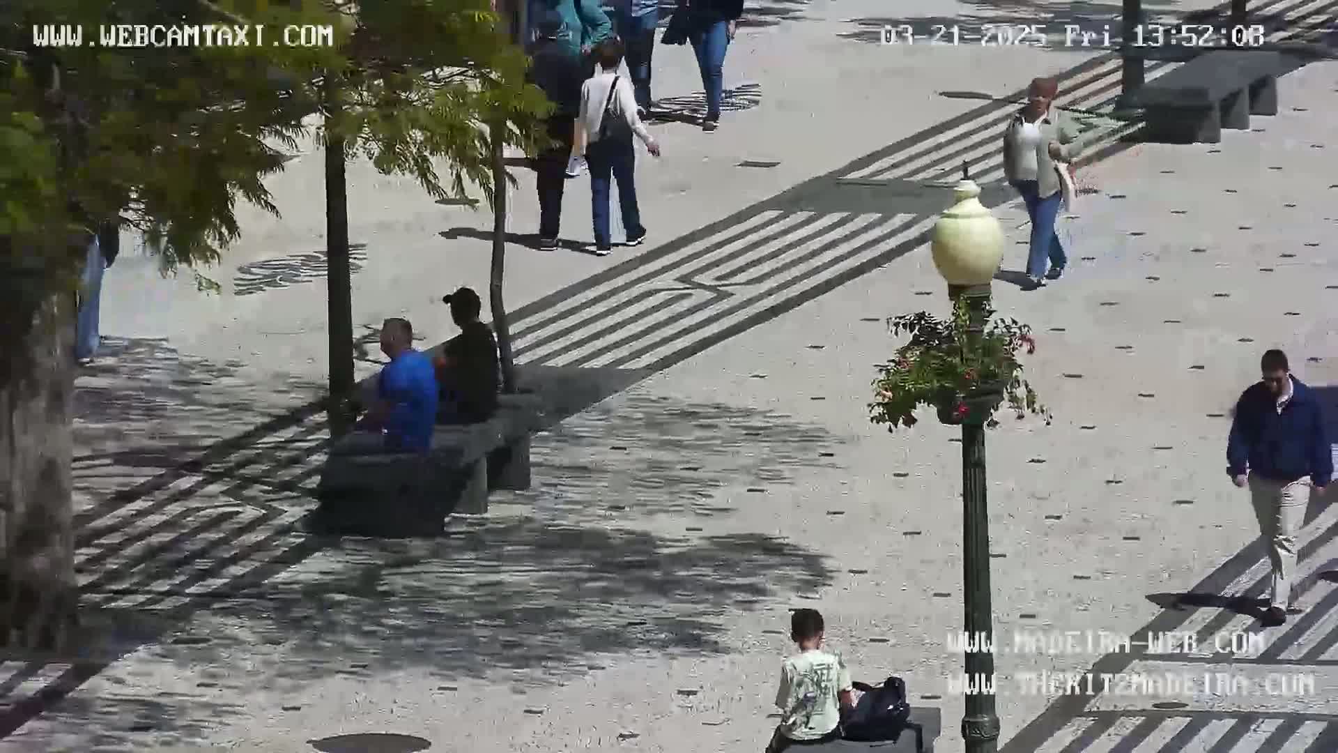 A sunny outdoor plaza features several people walking and sitting on benches, with patterned pavement and a lamppost.