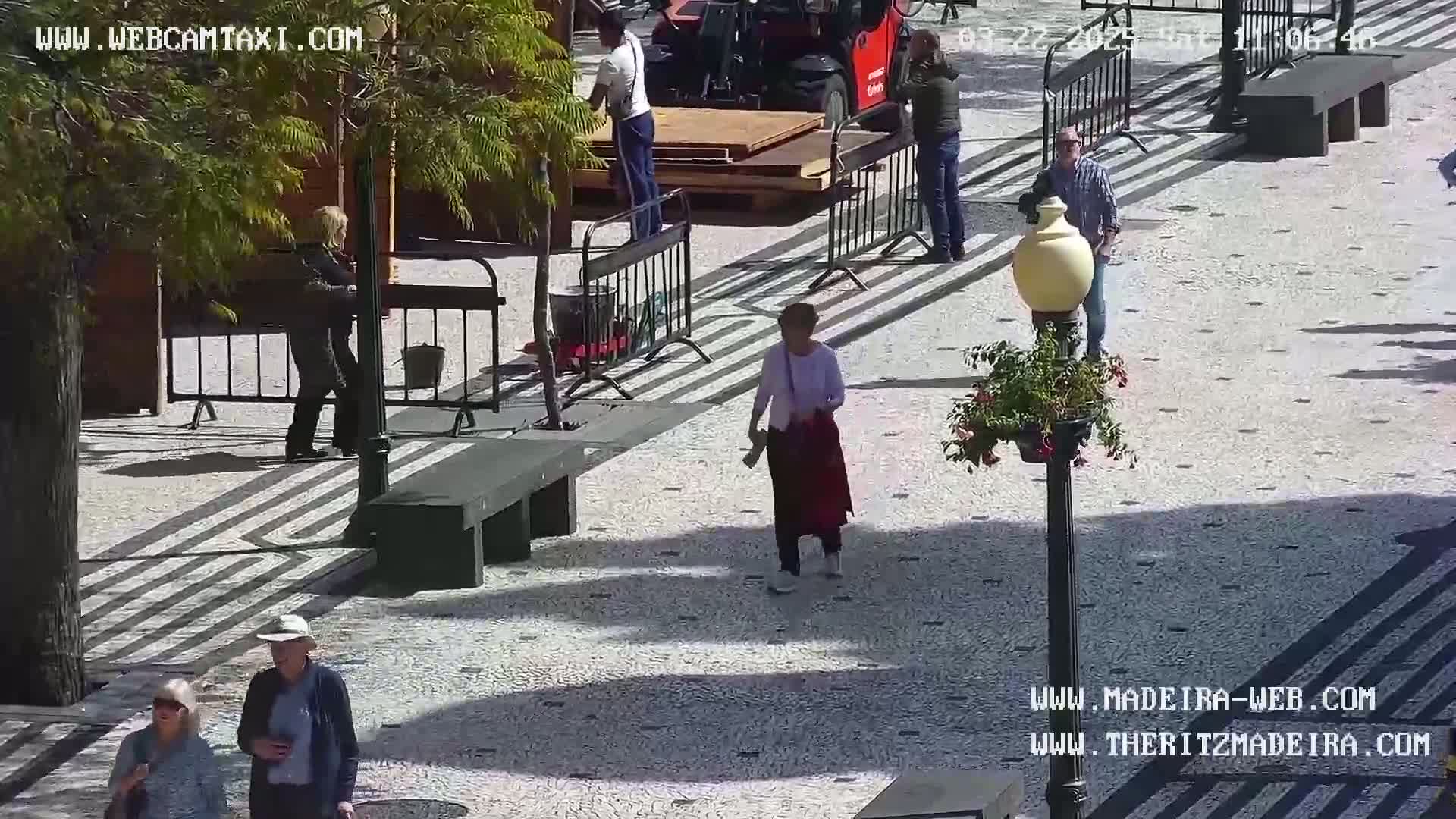 A sunny outdoor plaza scene shows several people walking amidst benches, barriers, and a lamppost, with construction work occurring in the background.