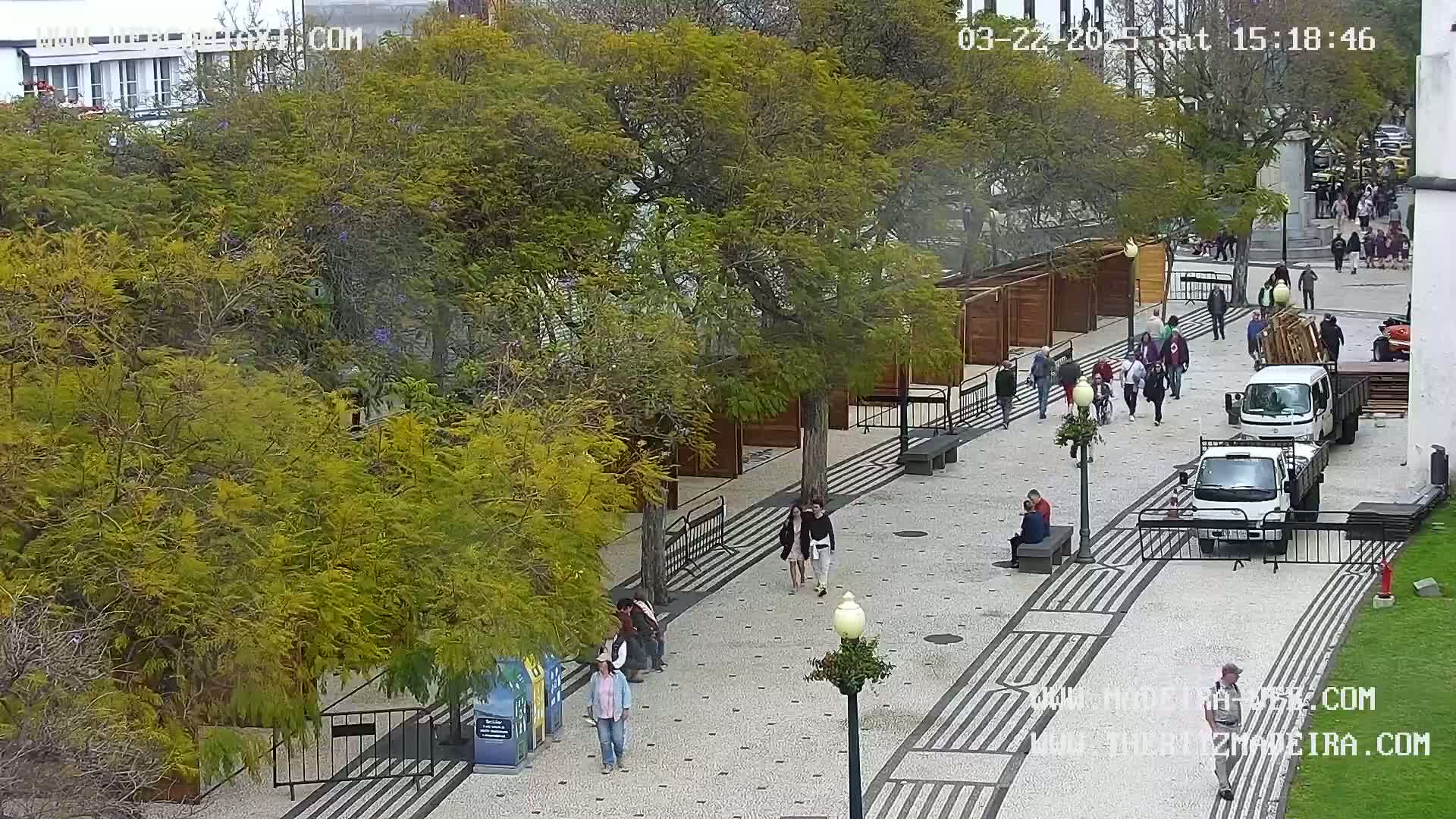 A paved pedestrian area with wooden stalls and several people walking, under an overcast sky, surrounded by lush green trees.