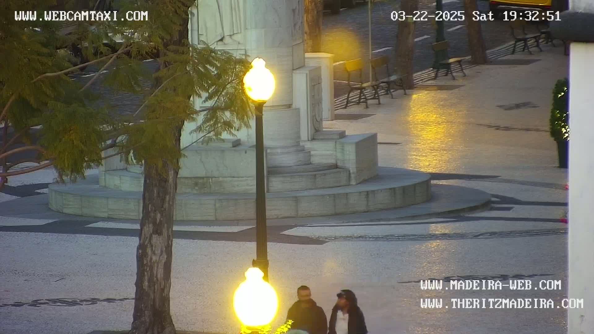 A stone monument with steps is in a plaza at dusk, illuminated by a lamppost and streetlights, with two people walking nearby.