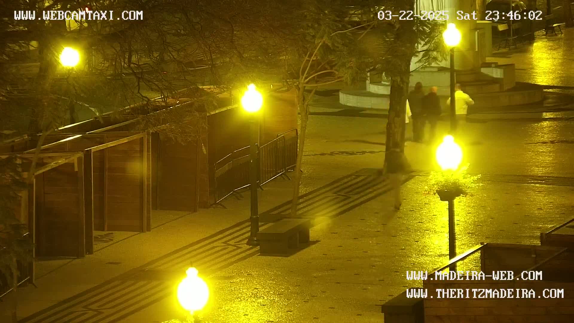 A wet, nighttime outdoor scene shows a paved plaza with several lampposts casting a yellow glow, some wooden structures, and a few people walking in the distance.