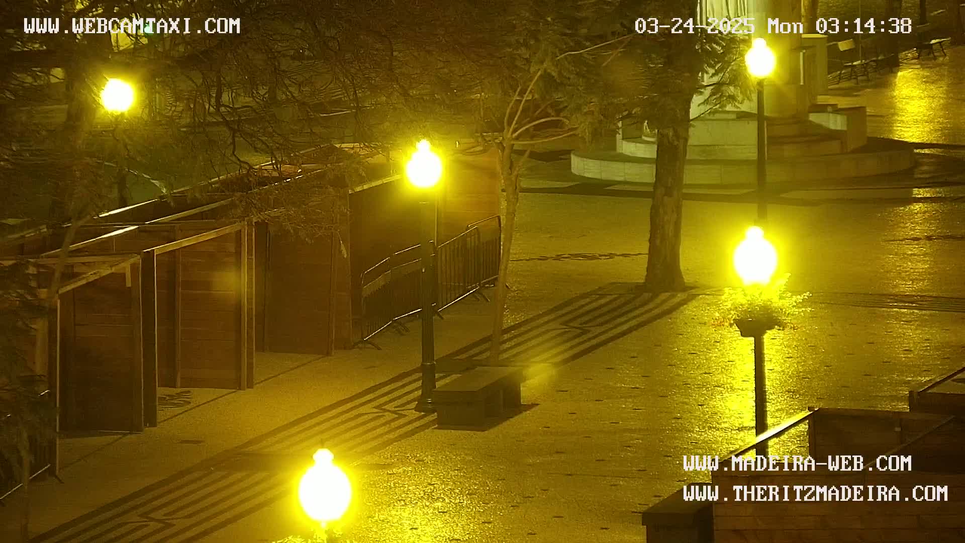 A wet, paved plaza at night is illuminated by several bright yellow streetlights, with several wooden structures and a metal barrier visible in the foreground.