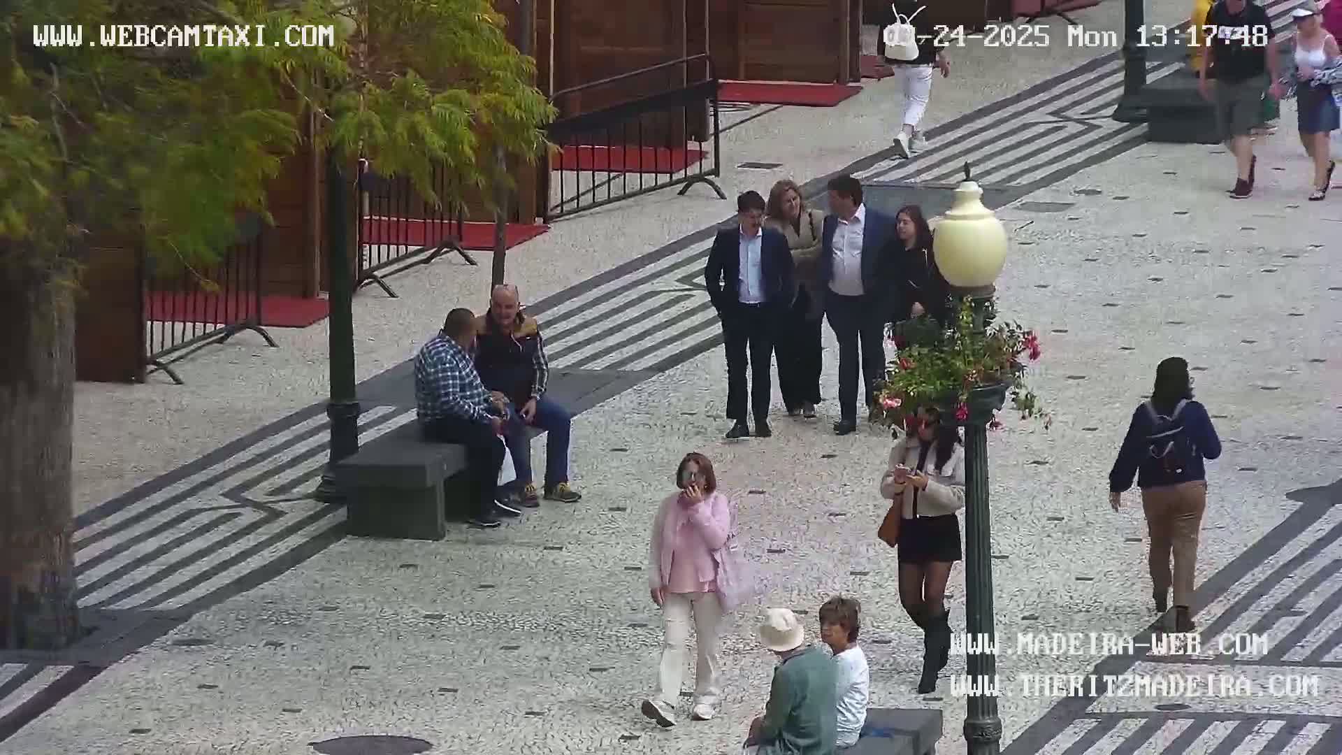 A paved outdoor plaza with several people walking and sitting, under a mostly cloudy sky.