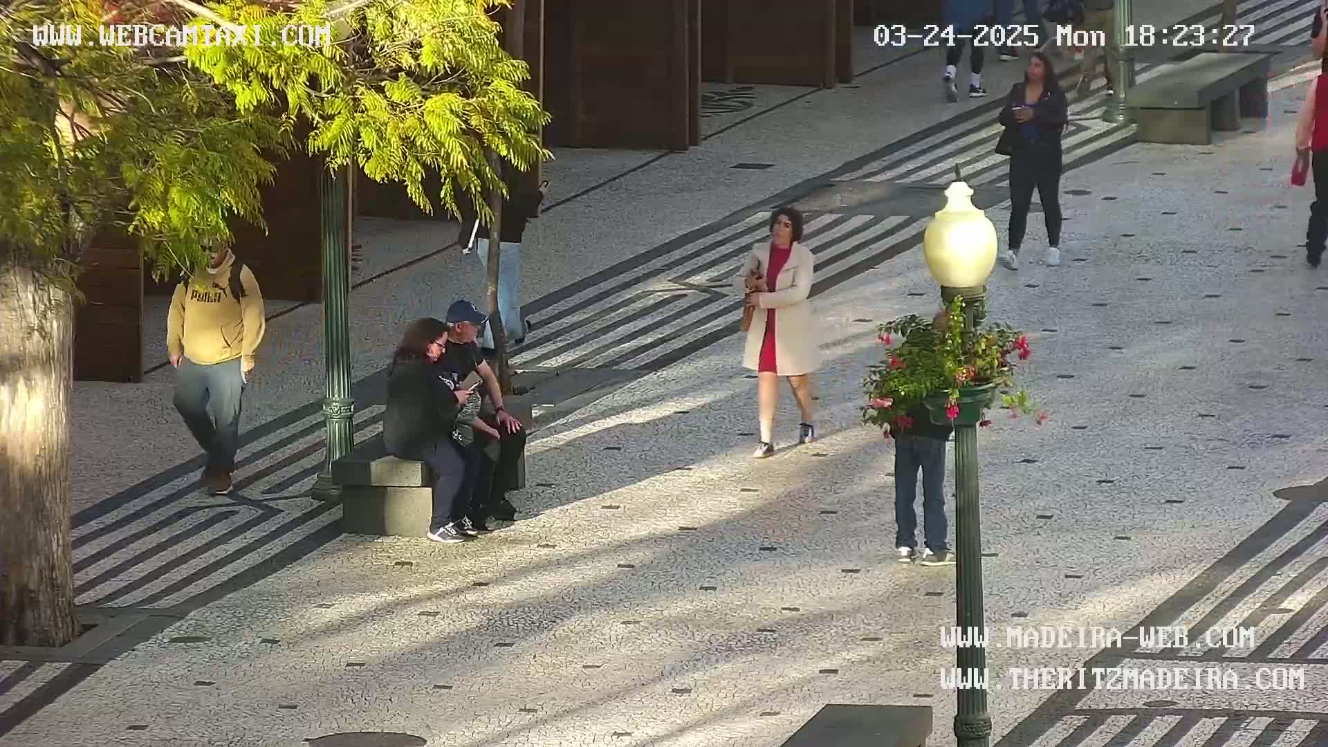 A paved outdoor area with several people walking and sitting, under a sunny sky.