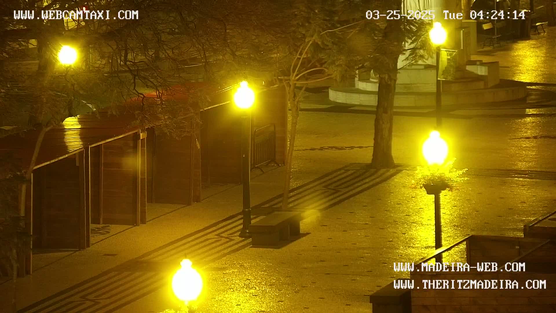 A wet, nighttime scene shows several lampposts illuminating a paved plaza with a row of dark wooden structures, and a tree casts shadows on the ground.