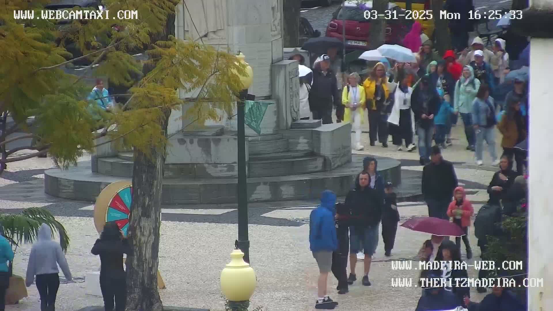 A rainy day scene in a plaza shows many people walking under umbrellas around a stone monument and a large wheel game.