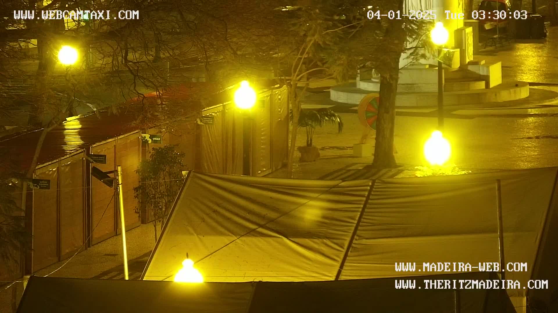 A nighttime scene of a wet paved area with several structures, including what appears to be temporary stalls or tents, illuminated by bright yellow streetlights.