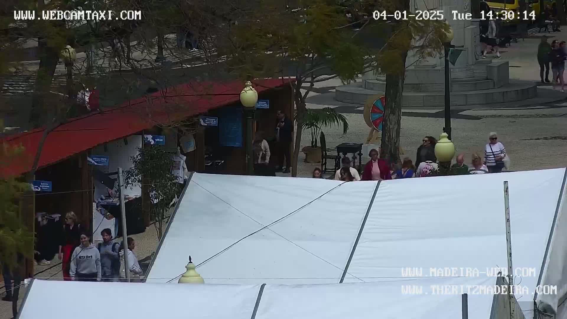 A fair or market scene outdoors on a partly sunny day, featuring several people walking around market stalls and tents.