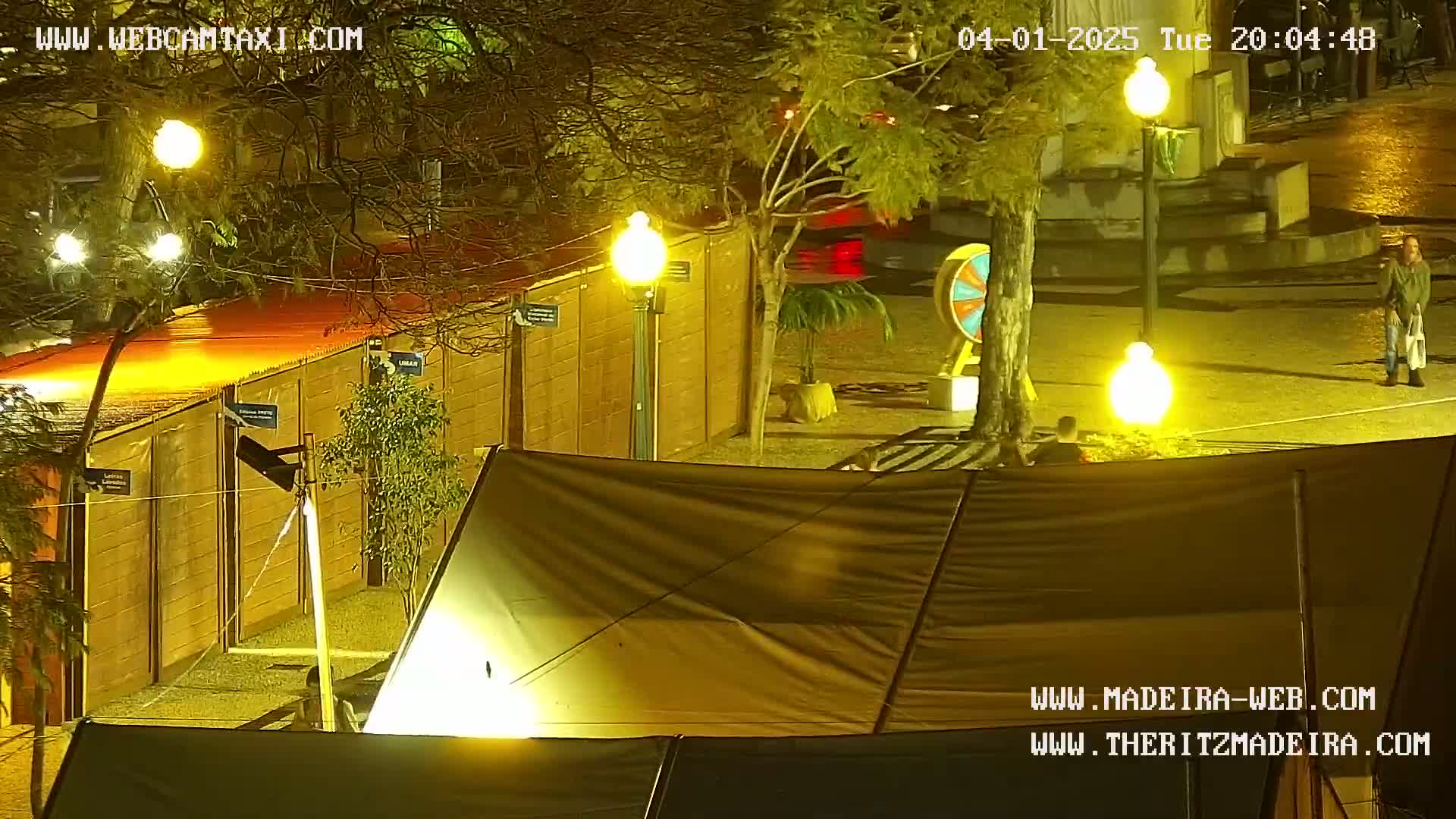 A nighttime outdoor scene shows a fenced-in area with several tents, a large wheel, and streetlights illuminating a wet paved area.