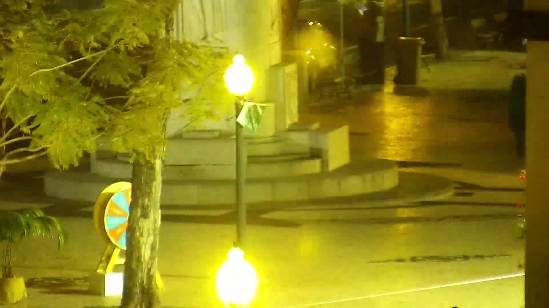 A wet, nighttime scene shows a plaza with a stone monument, a spinning wheel, trees, and illuminated lampposts.