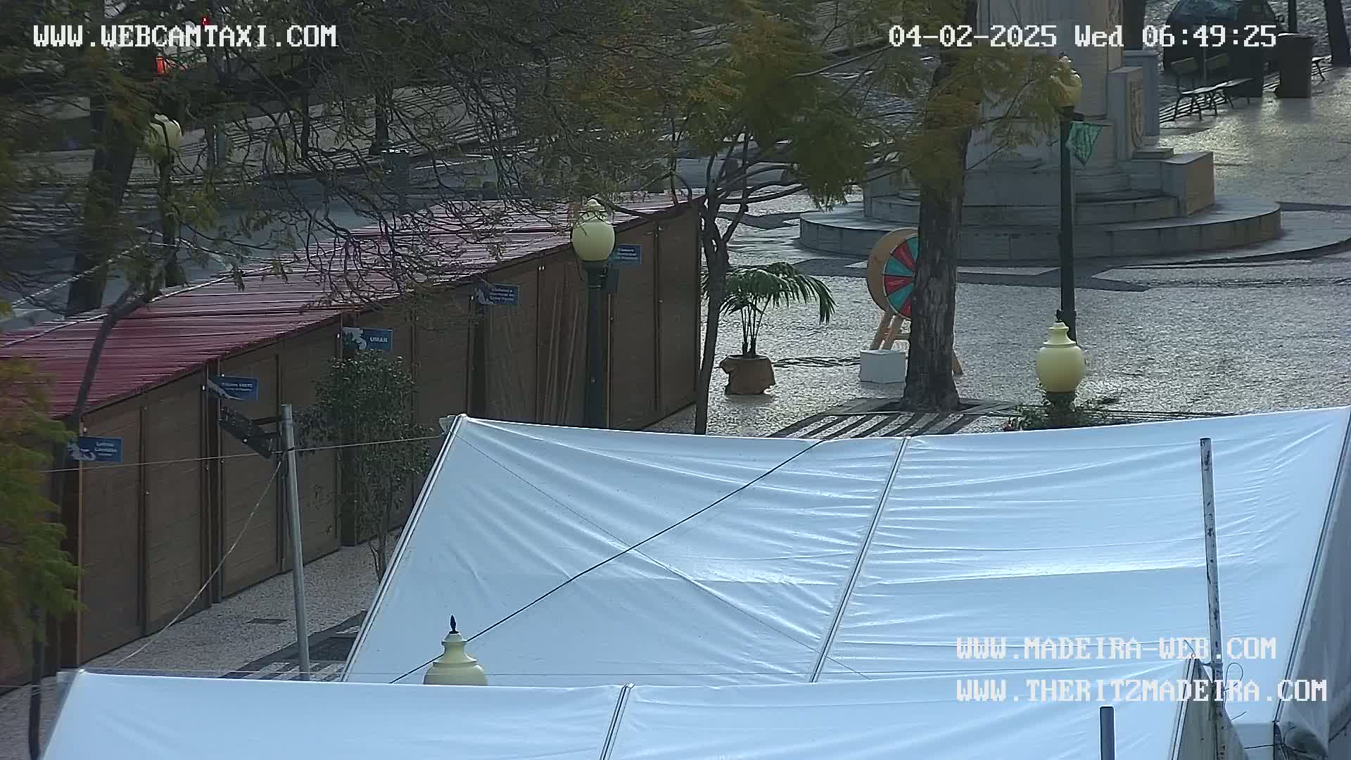 A view of a plaza with several wooden structures, a large white tent, and a small potted plant, under overcast conditions.