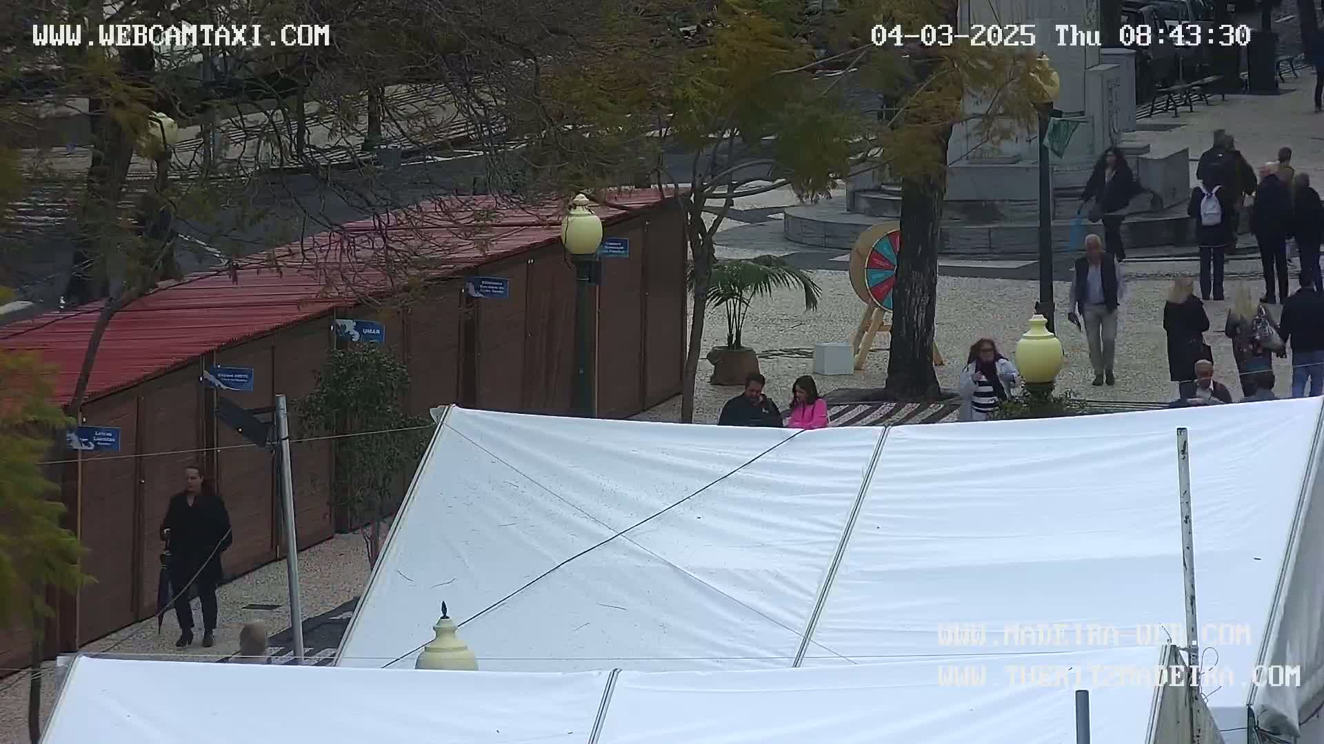 An outdoor scene on a partly cloudy day shows several people walking around a paved area with temporary stalls and tents, and a large wheel.