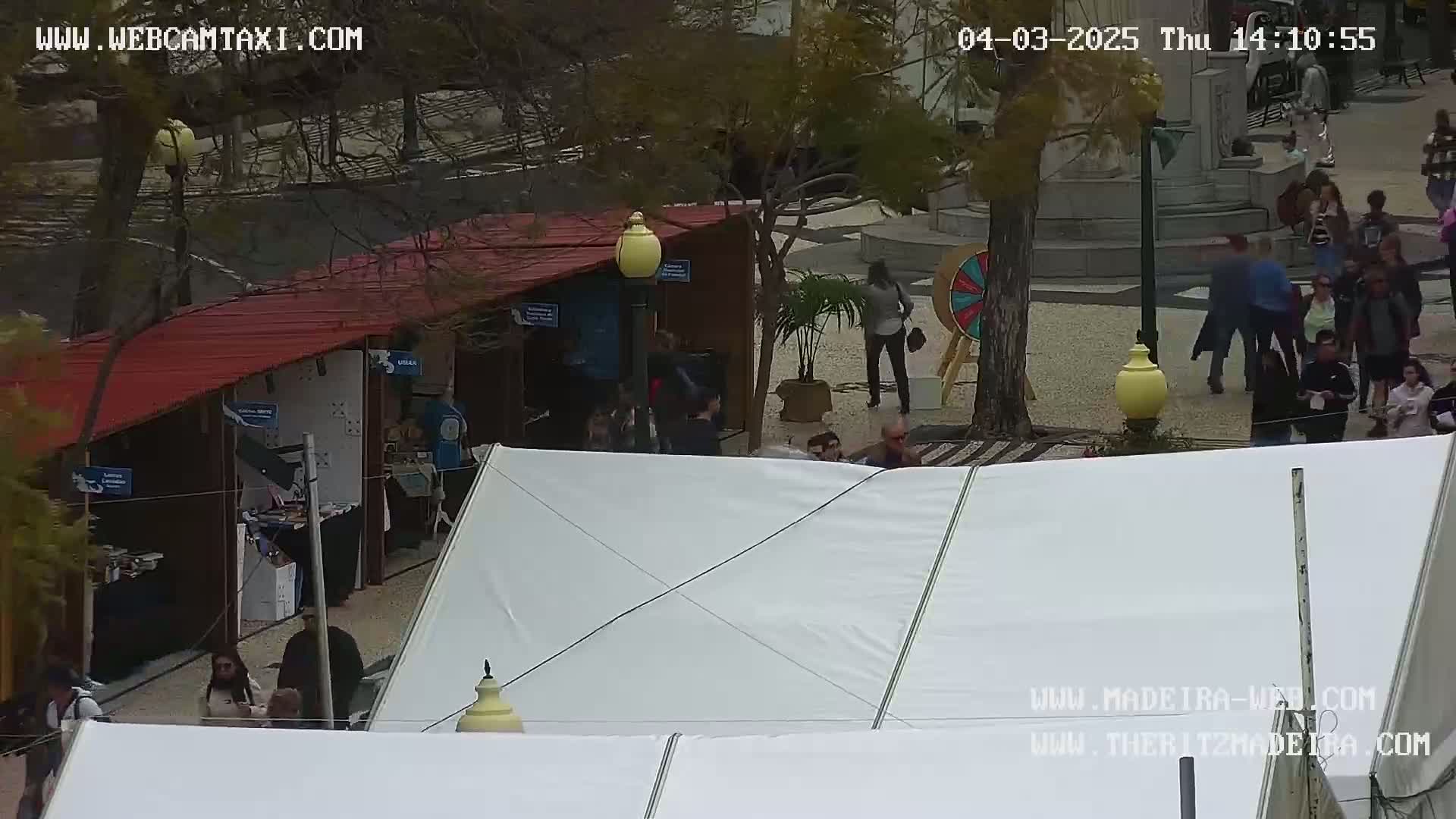 An outdoor market scene with several people milling about under an overcast sky, featuring various stalls and a large white tent.