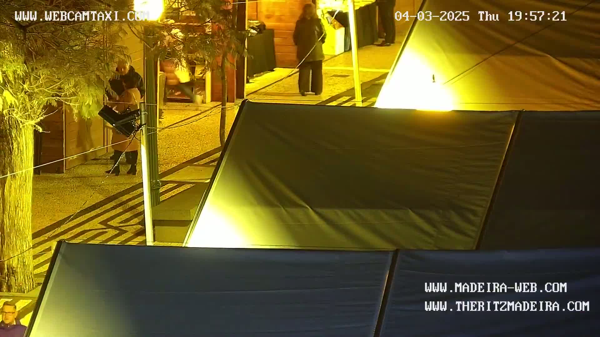 At night, under bright artificial lights, people stand near several dark-colored tents set up outdoors on a paved area.