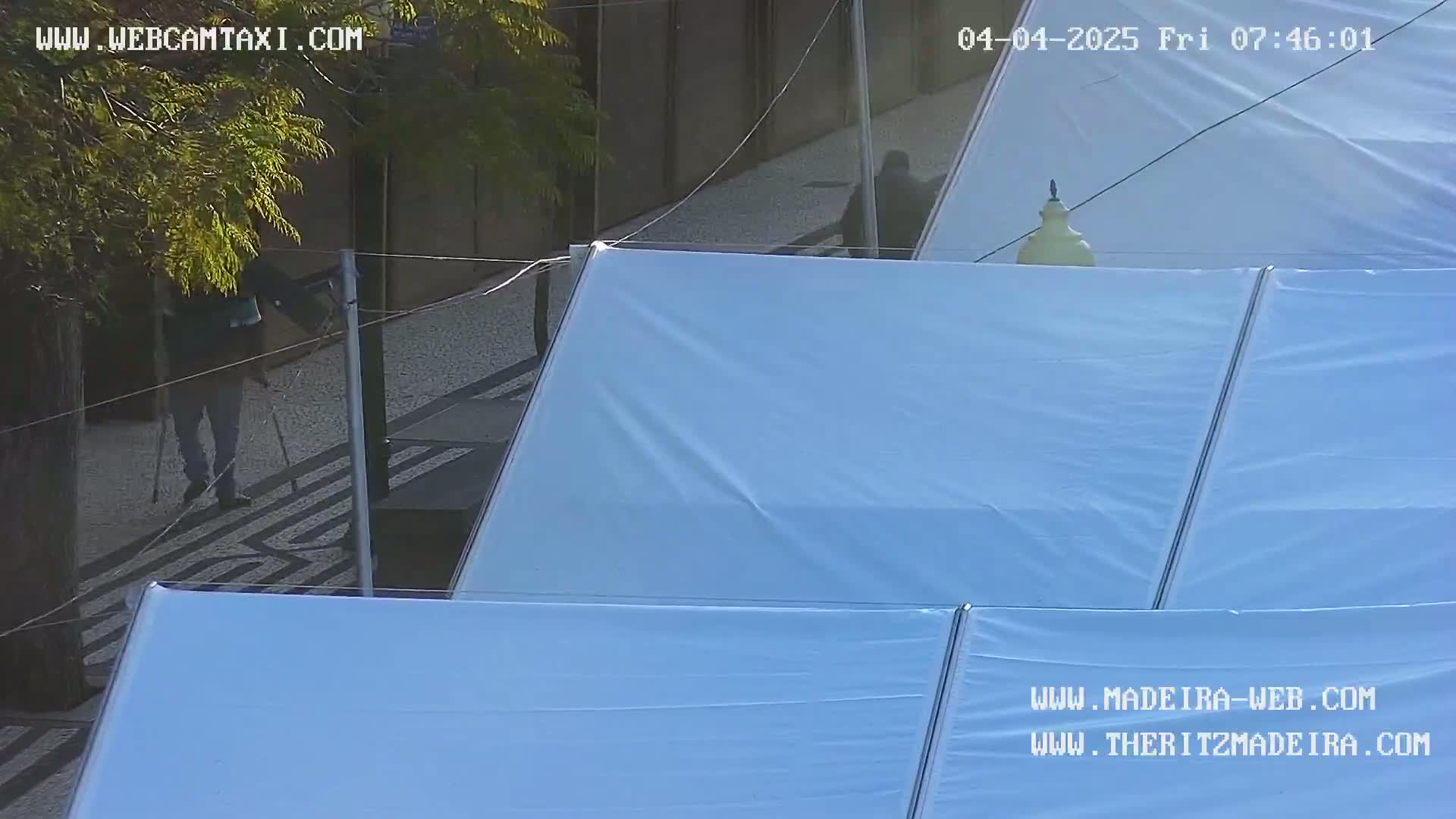 A person walks on a sidewalk next to a building partially covered by large white fabric sheets under a sunny sky.
