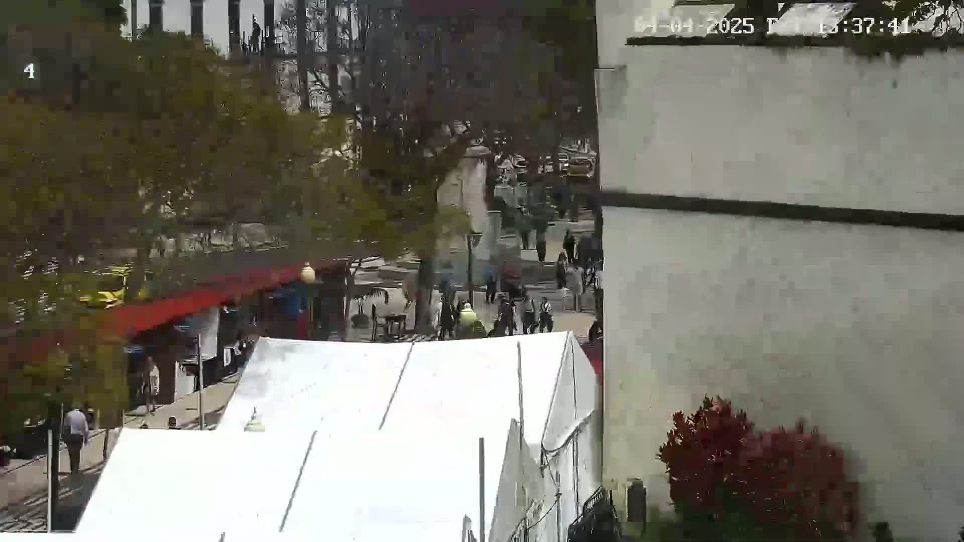 A high-angle view shows a city street scene with people walking, some cars, trees, buildings, and white tents on a partly sunny day.