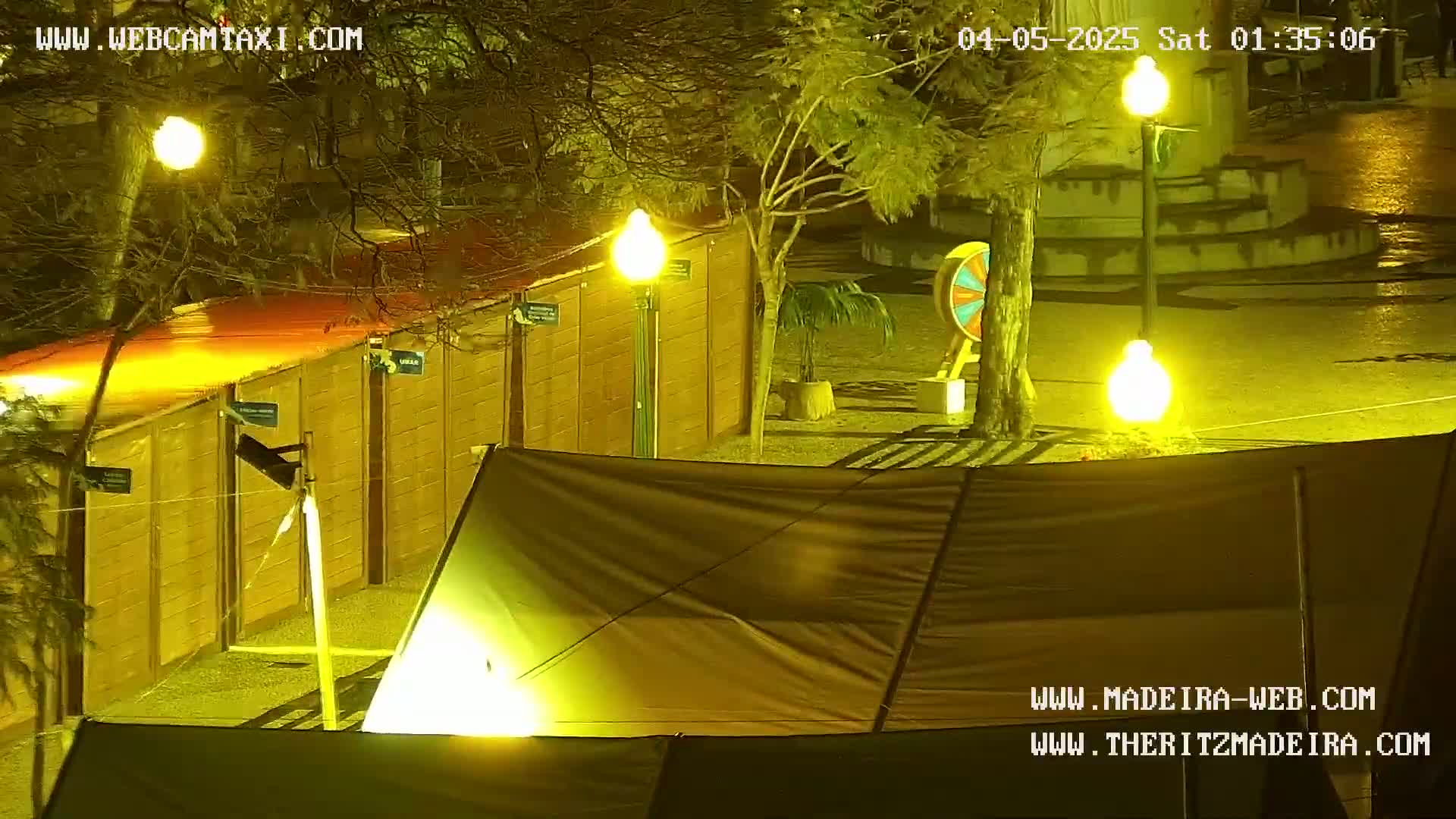 A nighttime view of a fenced-in area with a tent, illuminated by streetlights, showing some wet pavement.