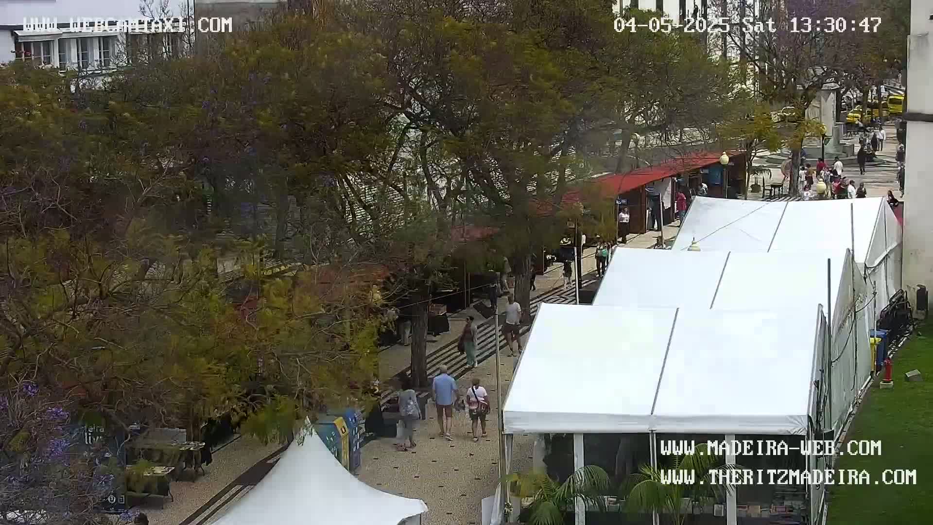 An outdoor plaza scene on a partly sunny day shows several people walking among trees, small market stalls, and several large white tents.