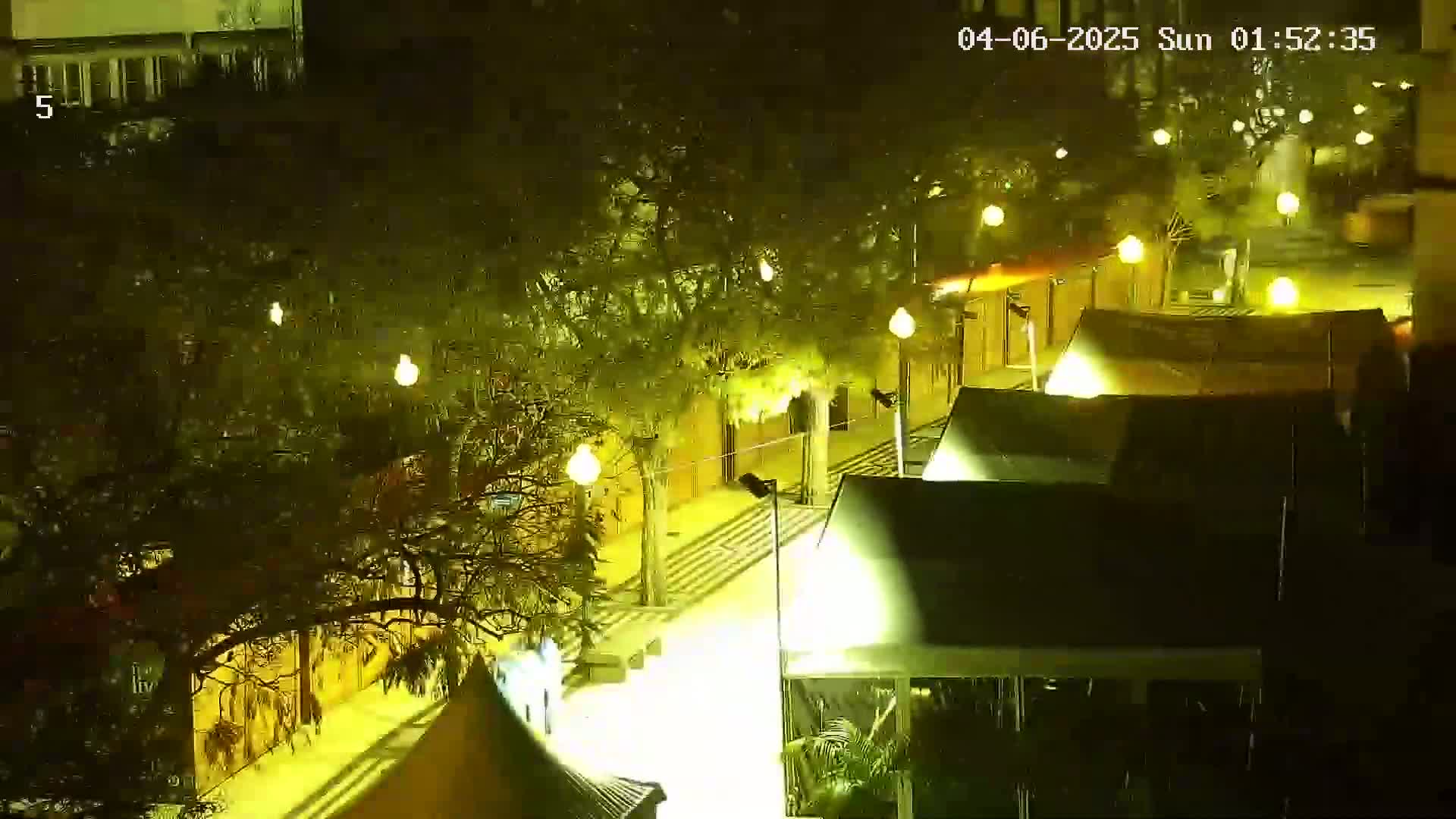 A nighttime, illuminated outdoor scene shows a paved area with several dark-colored stalls or tents under trees, lit by numerous yellow lights.