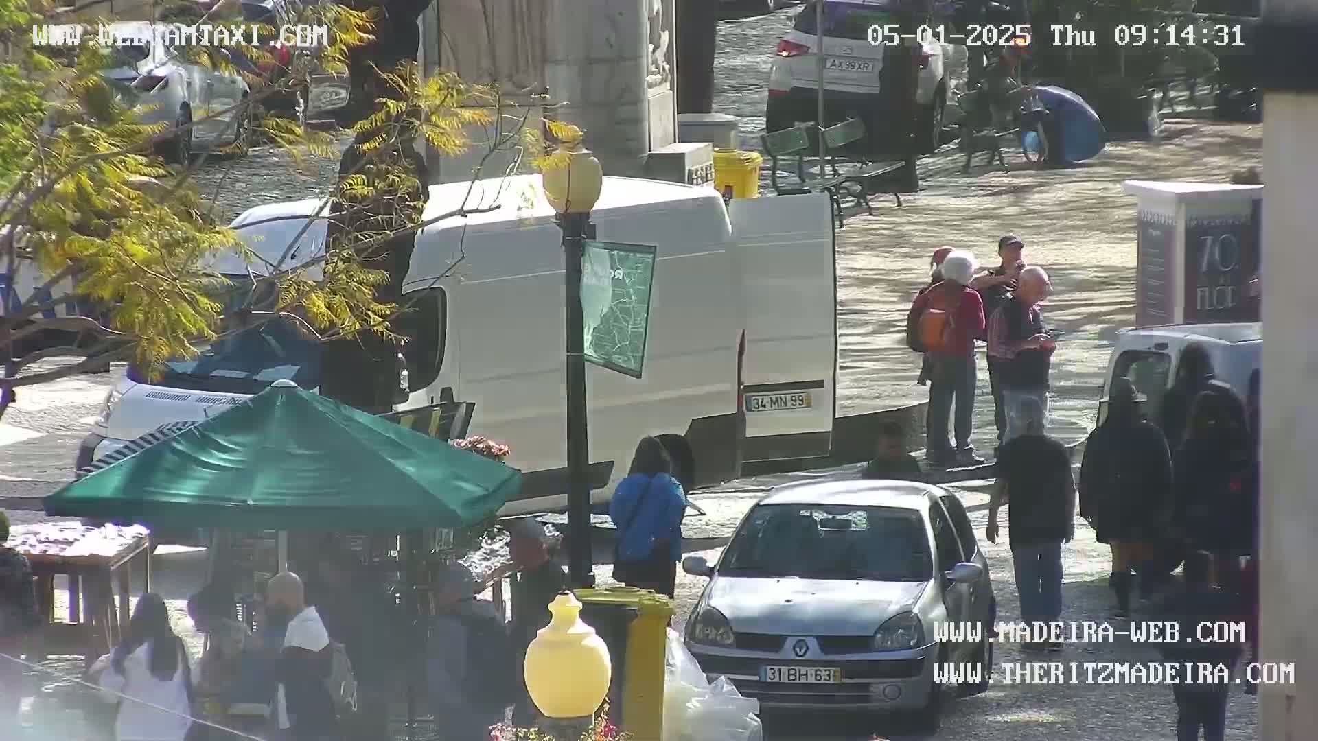 A sunny outdoor scene shows a plaza with several people, parked cars, a white van, and a green umbrella covering a small stand.