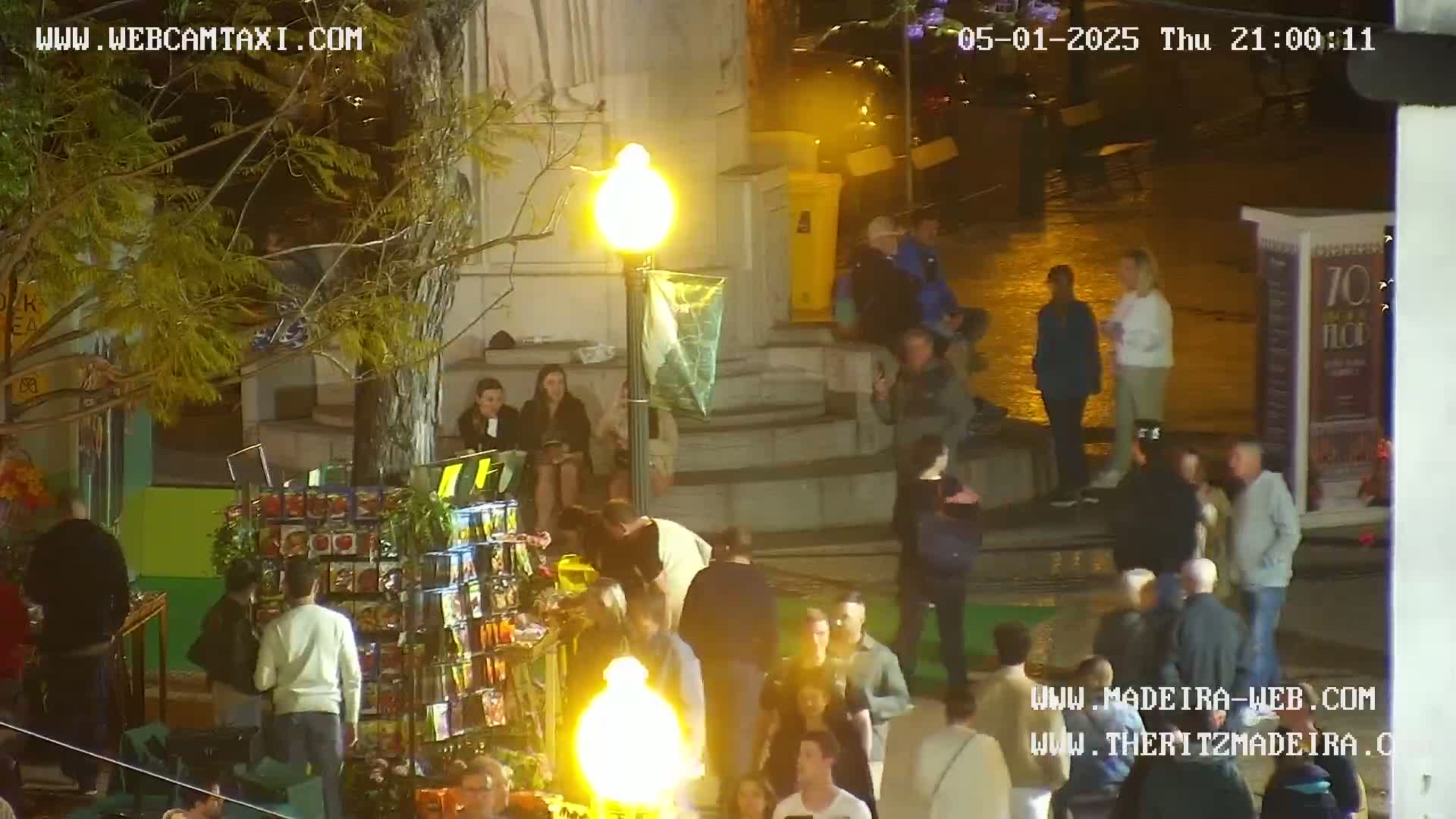 A nighttime outdoor scene shows a crowd of people gathered around a small vendor's stall, under a lamppost, on a wet street.