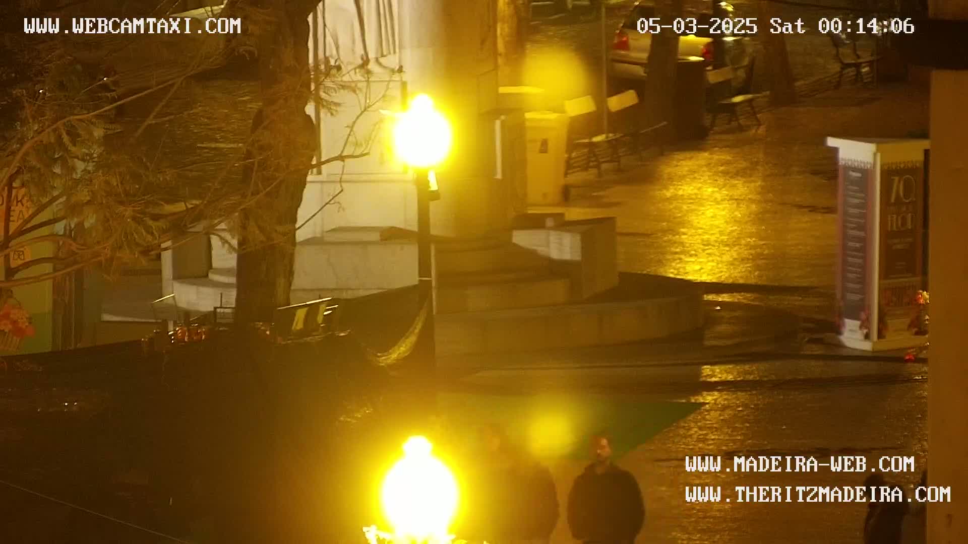 A nighttime, wet city scene shows two figures standing near bright yellow streetlights, with a dark, possibly tree-covered area and buildings in the background.