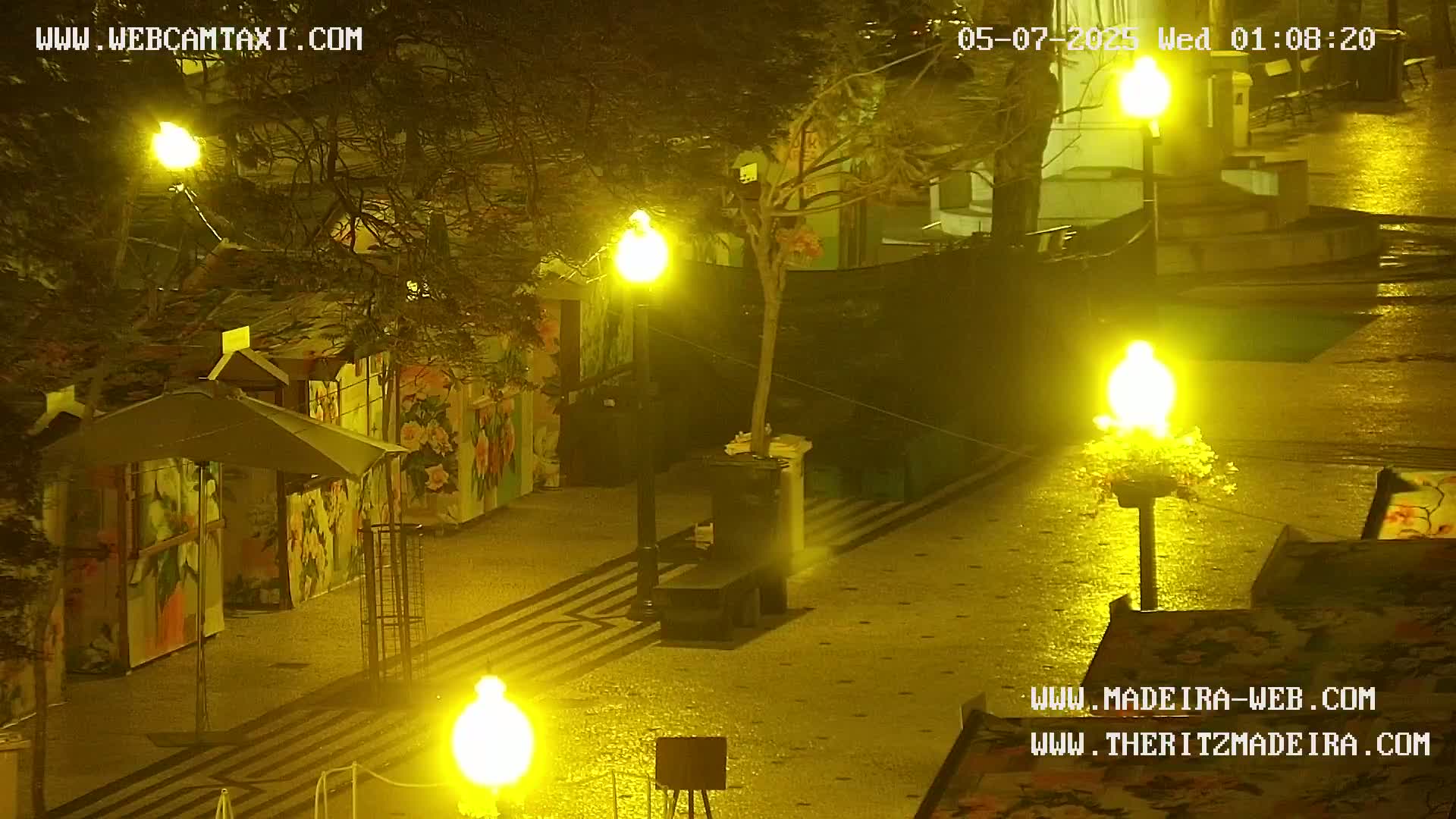A nighttime scene shows a paved area with several brightly lit lampposts, small stalls with colorful painted sides, and a few umbrellas.