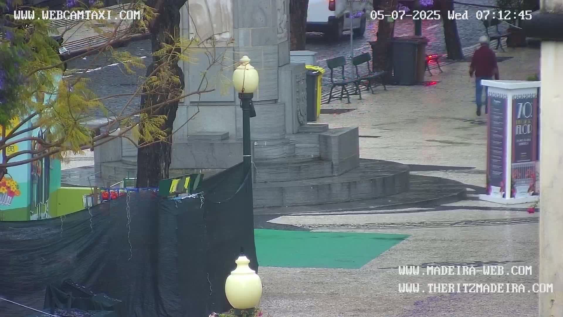A wet, outdoor plaza scene features a stone monument, benches, a person walking, and construction-related barriers and coverings.