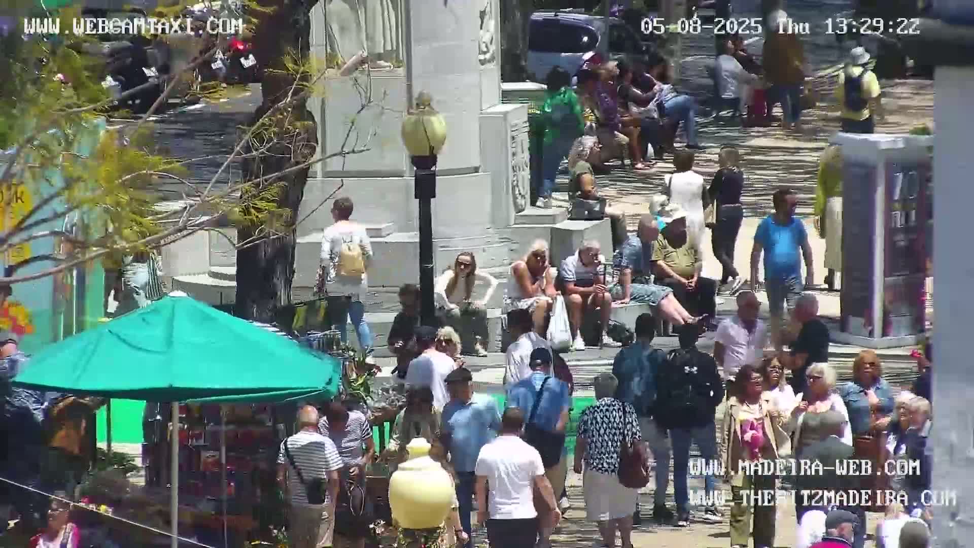 A sunny day shows many people gathered in a plaza around a monument, some sitting and others standing near a market stall under a green umbrella.