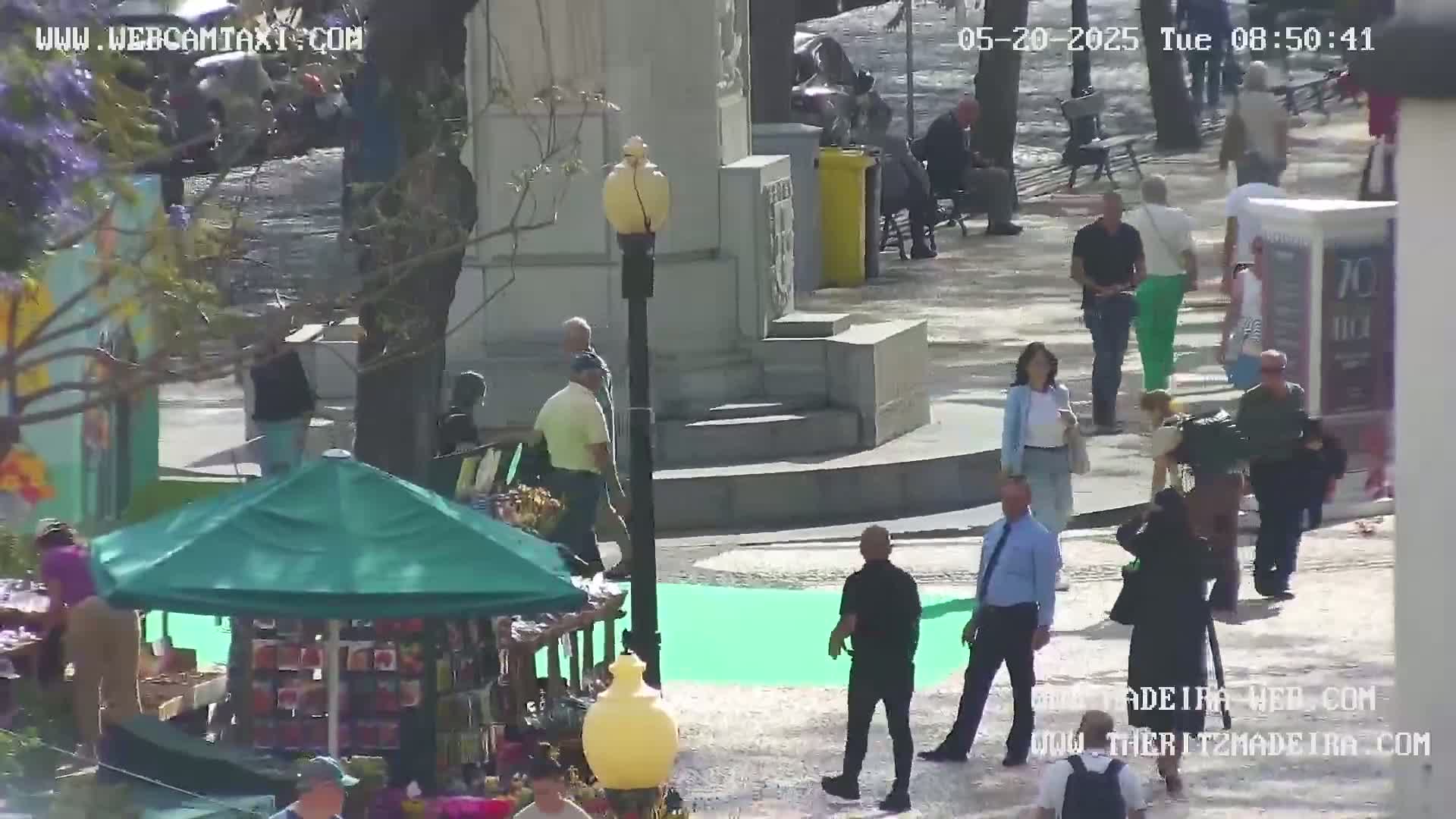 A sunny outdoor plaza scene shows people walking around a monument and a small market stall with a green umbrella.
