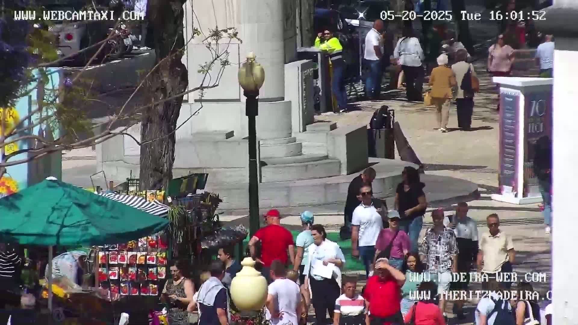 A sunny outdoor plaza scene shows a crowd of people walking near a street vendor under a green umbrella, next to a large stone structure.