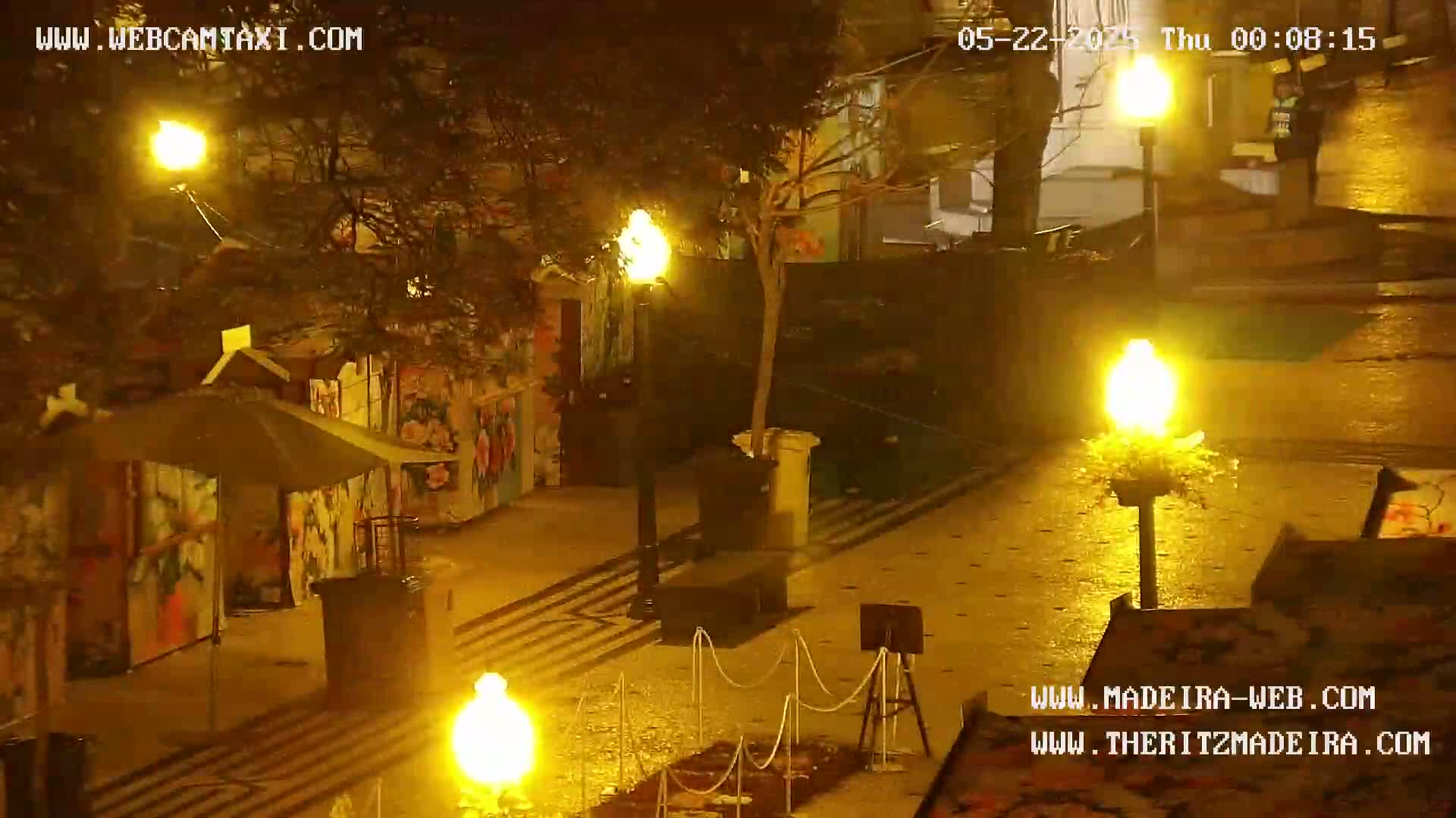A nighttime outdoor scene shows a paved area with several small structures, decorative lights, and a tree in planters.
